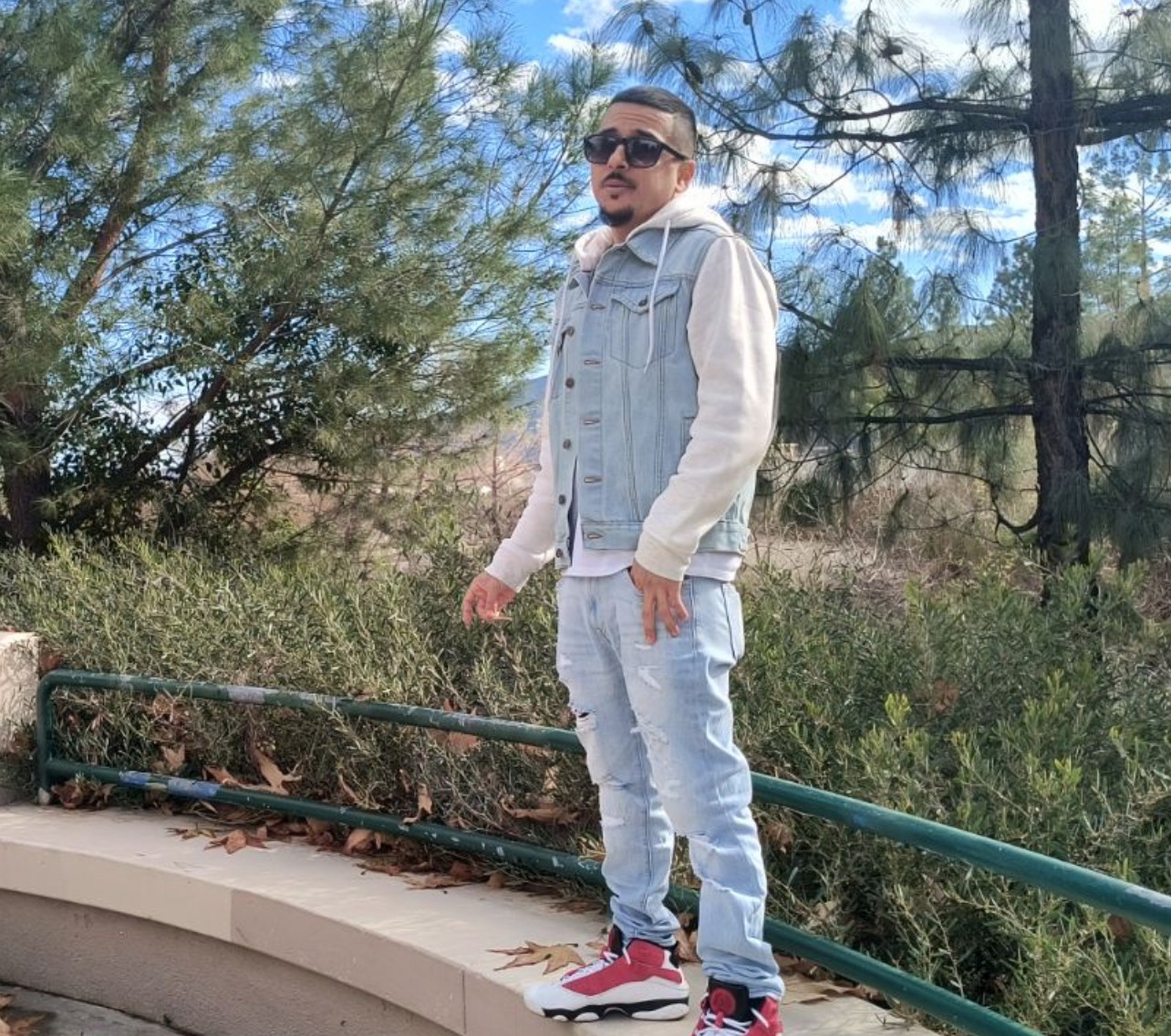 Person standing outdoors on a curved walkway with trees and a fence in background.