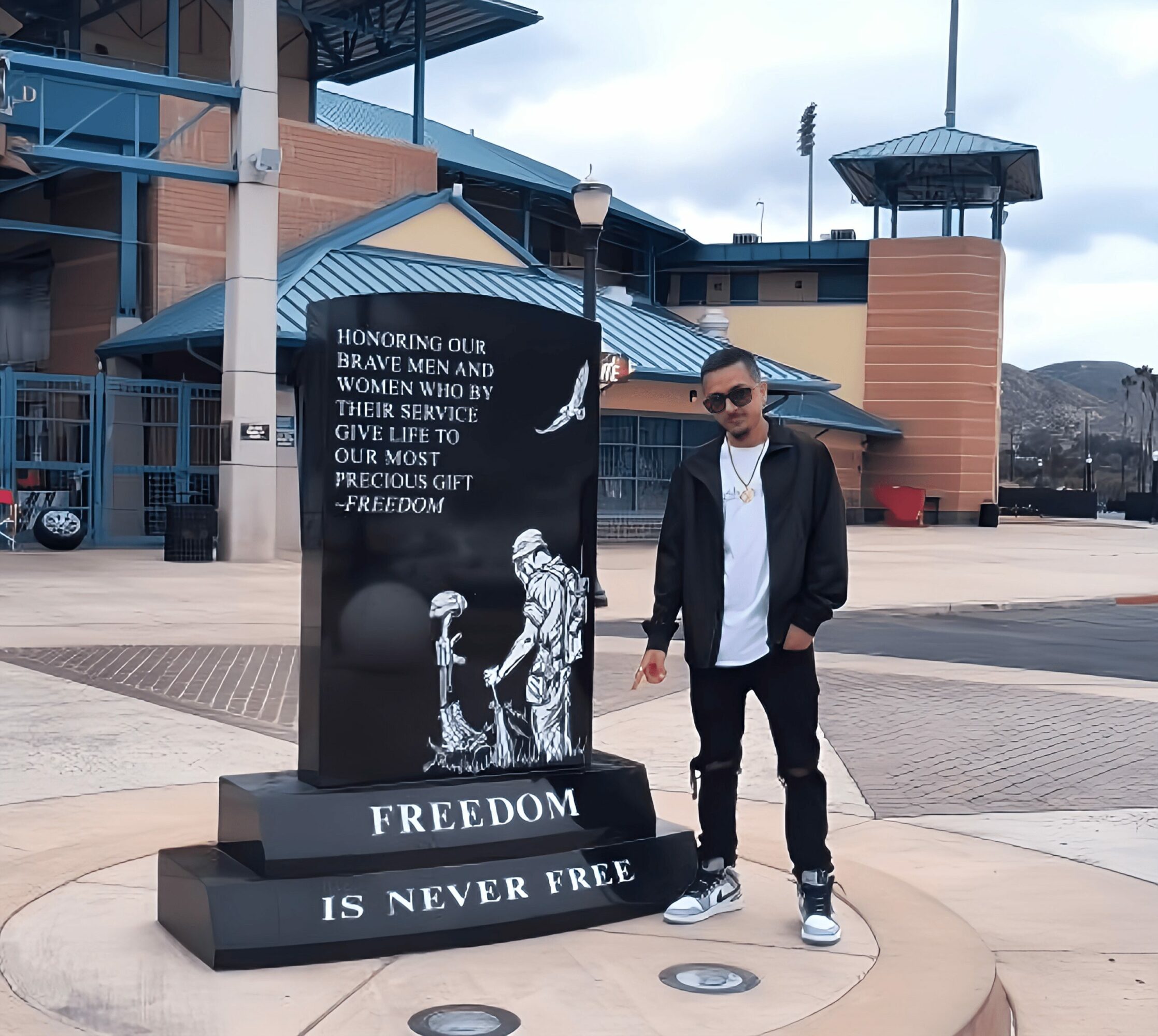 Young man in sunglasses and casual clothing standing next to a memorial plaque outdoors.