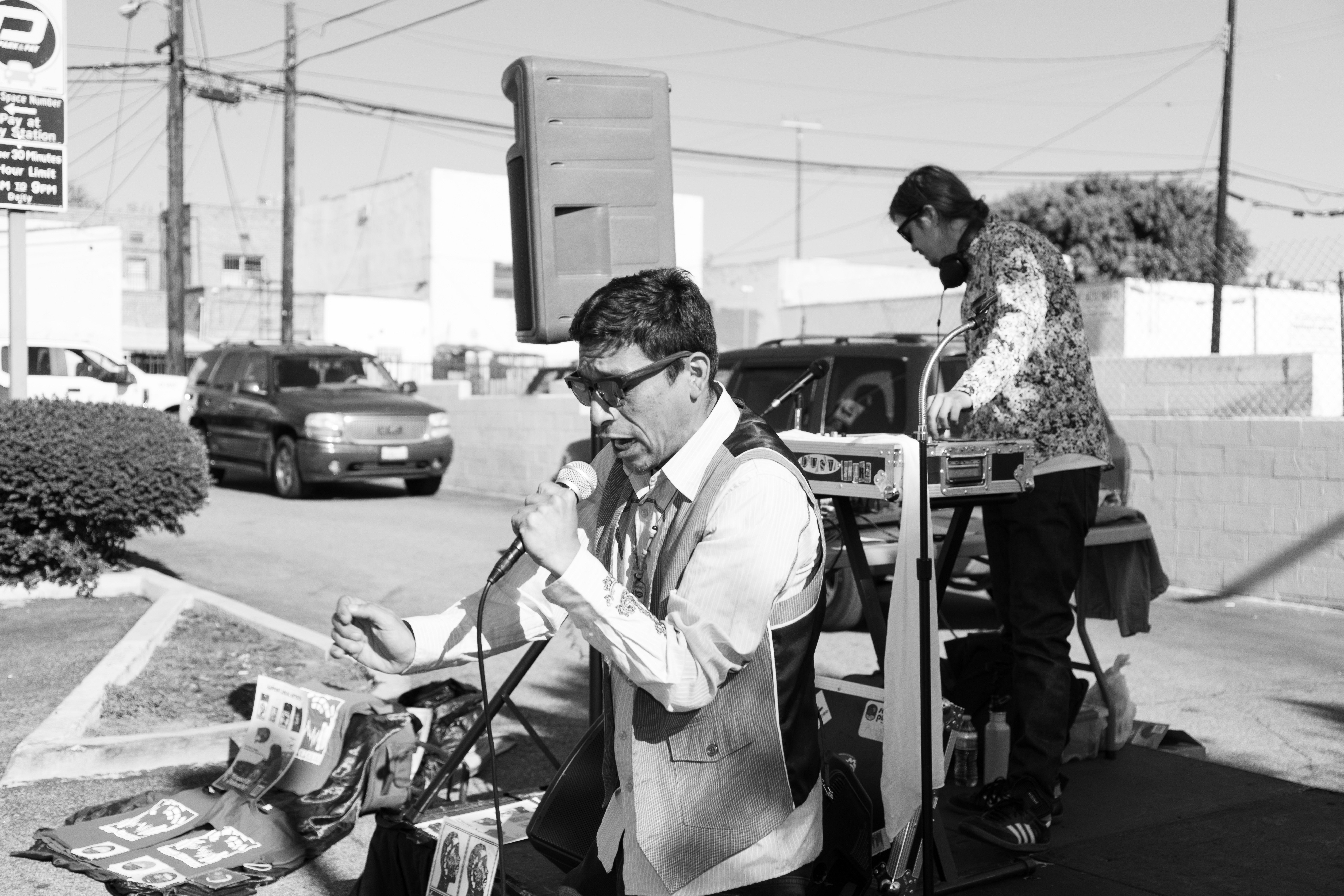 Man in glasses and vest using a public telephone booth outdoors, woman in patterned top in background, cars and buildings nearby.
