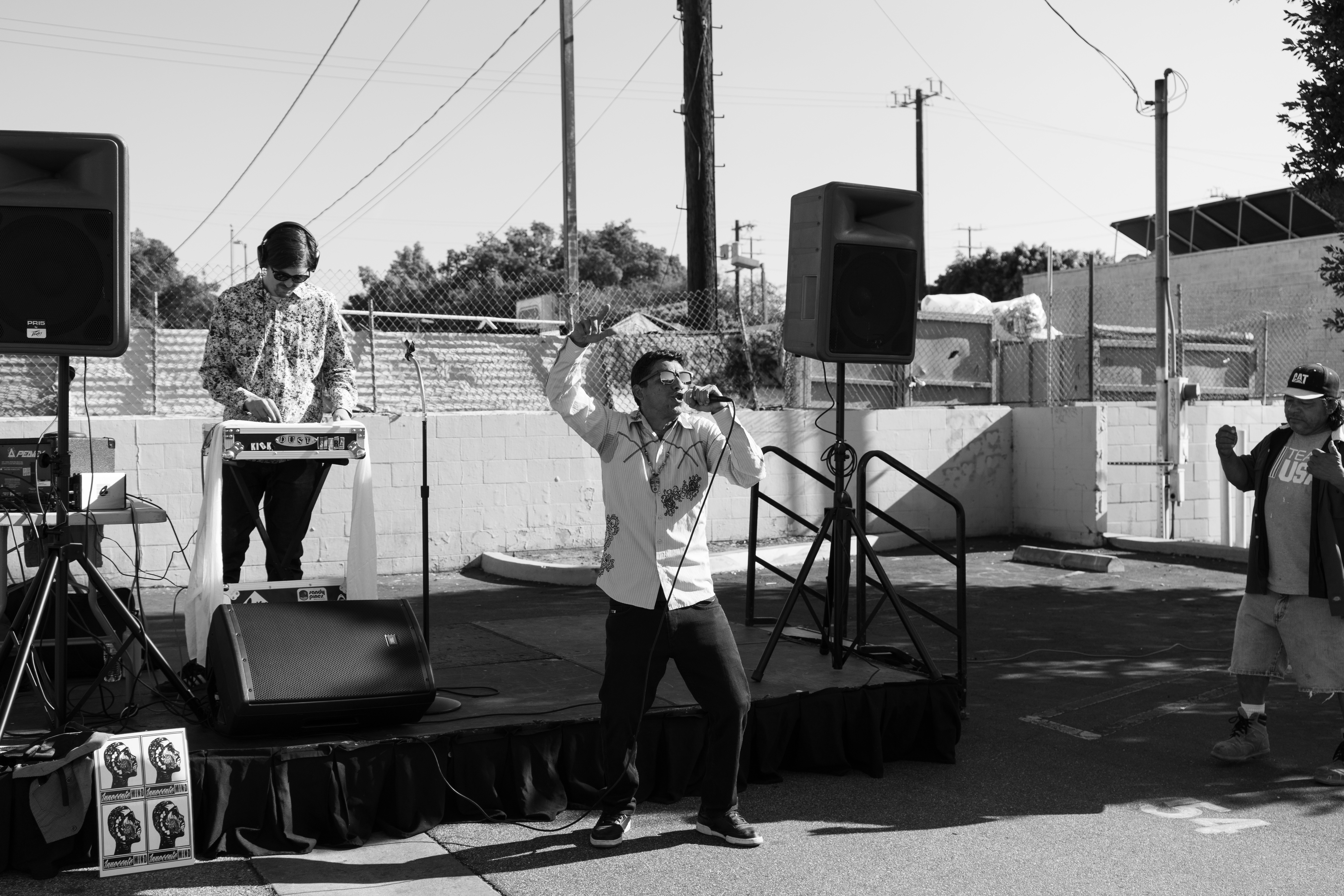 Person singing into microphone on outdoor stage with two other people nearby, sound equipment, and speakers.