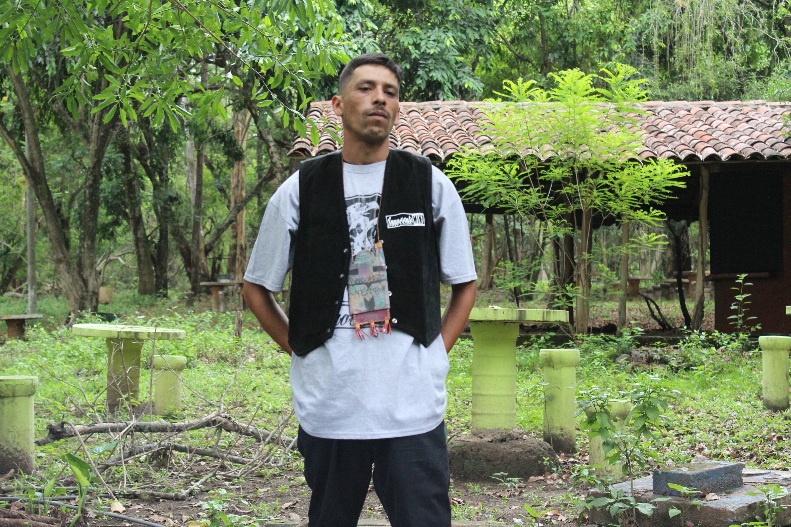 Young man standing outdoors with trees and greenery in background, wearing a white t-shirt and black vest, carrying a backpack.