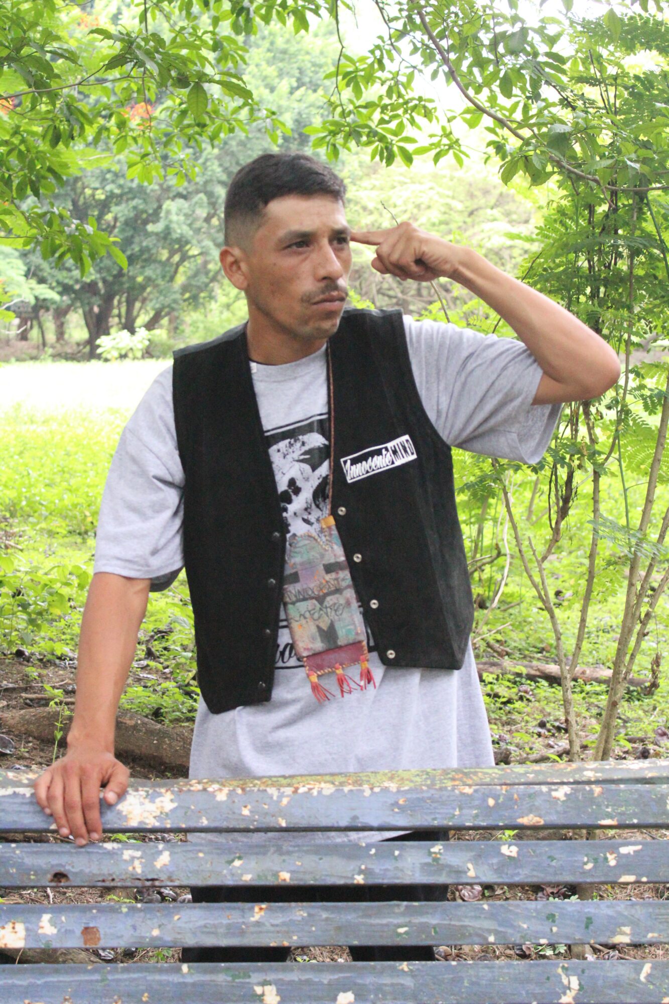 Man standing outdoors with trees and greenery, wearing a black vest over a gray T-shirt, holding a wooden bench.