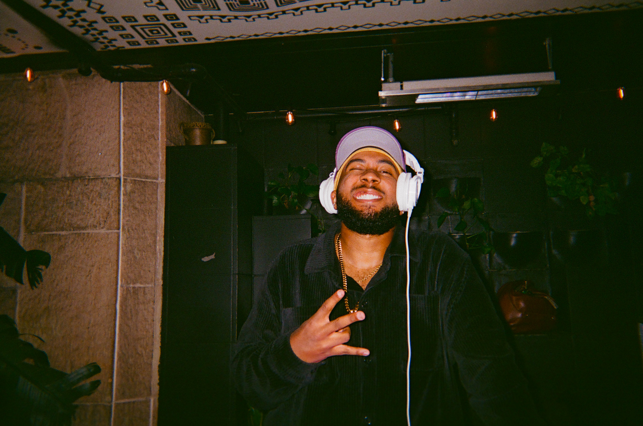Person wearing headphones and a hat, smiling and making a peace sign gesture indoors.