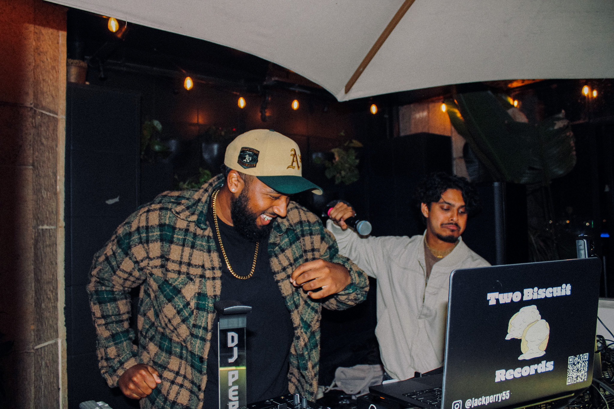 Two men standing at a music event, one wearing a plaid shirt and baseball cap, the other in a white shirt, with a laptop nearby.