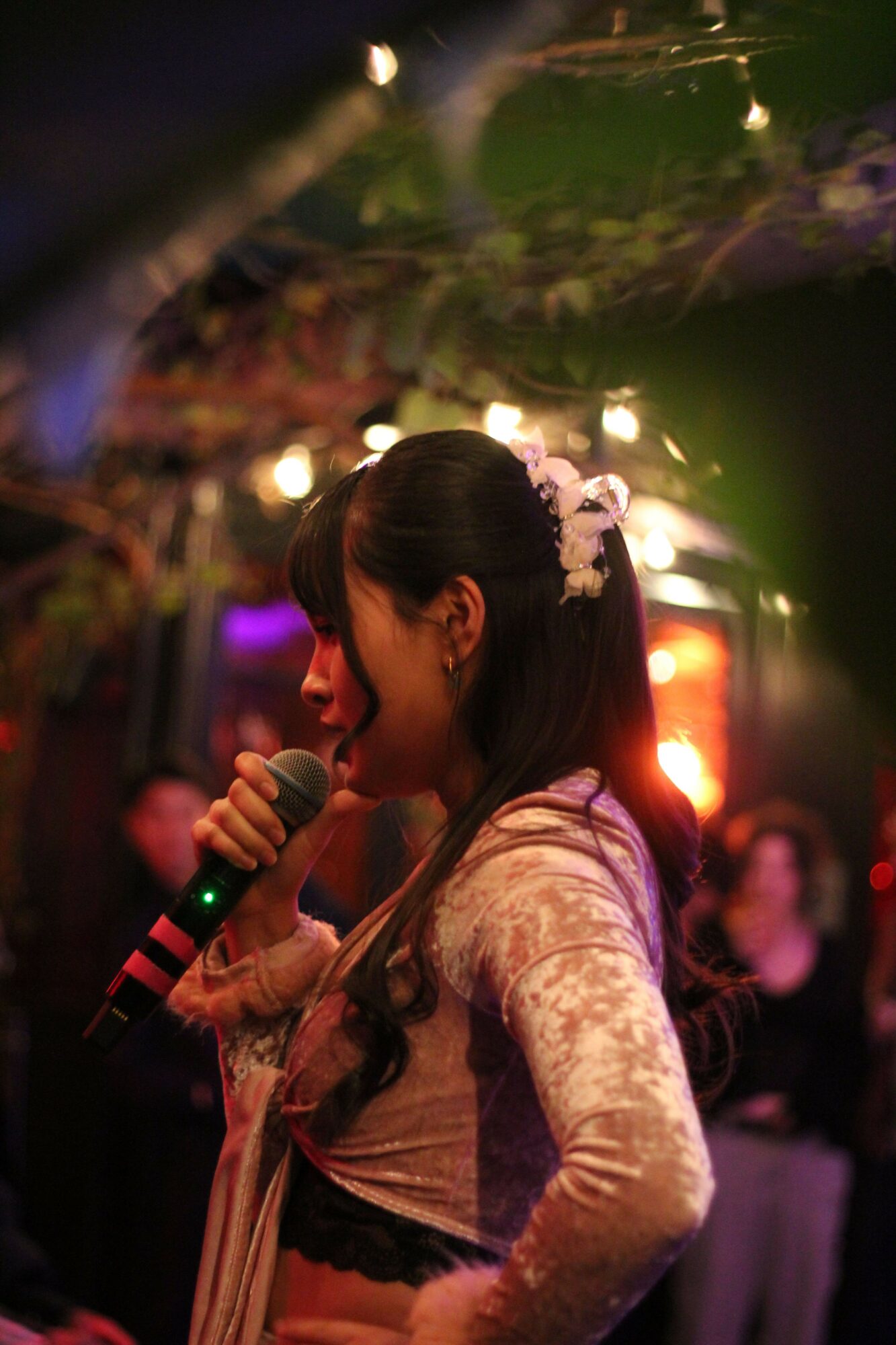 Young woman singing into a microphone at a lively indoor event with colorful lights and audience in background.