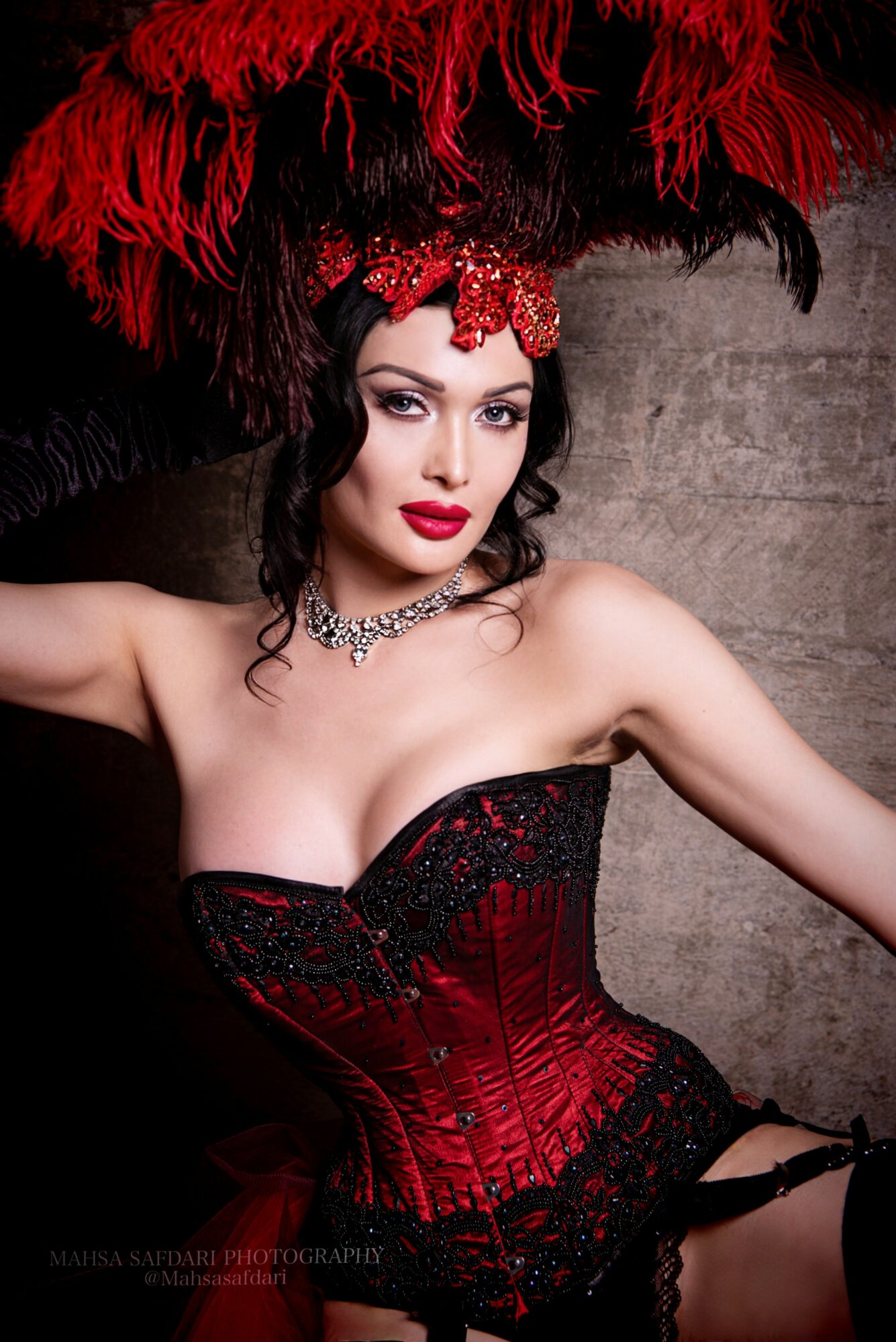 Woman in red and black costume with large feathered headdress, makeup, and jewelry, posing against a gray wall.