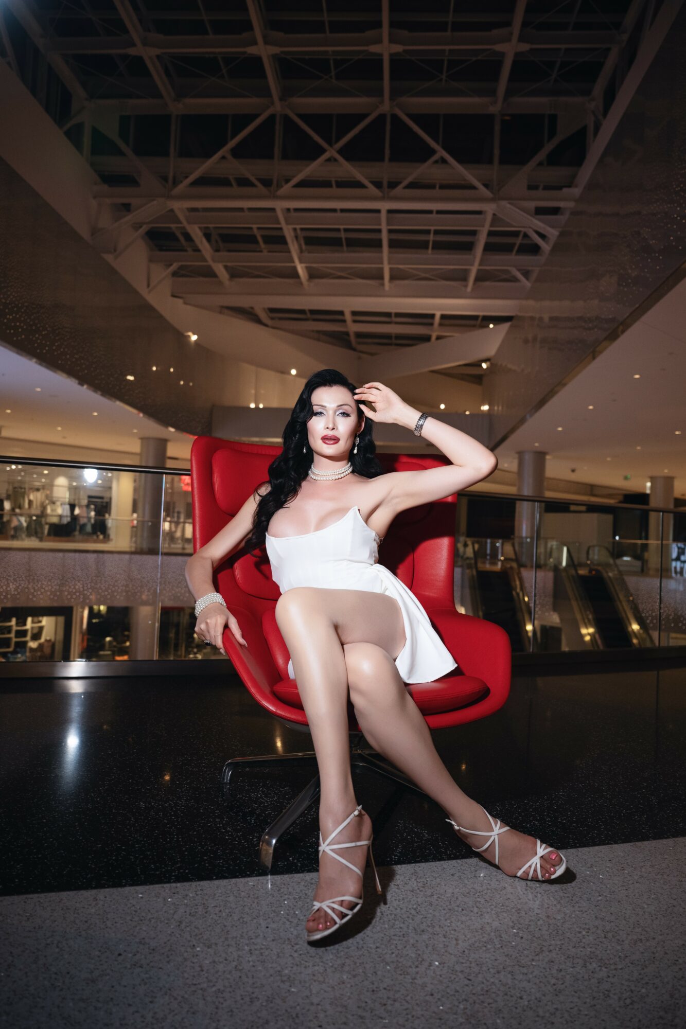 Woman with dark hair in white dress sitting on red chair in mall, ceiling structure above.