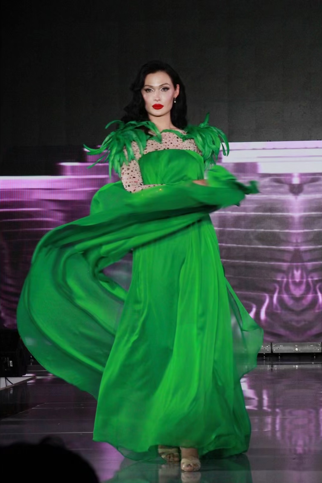Woman in a green gown with feathered shoulders, standing on stage with purple lighting in background.