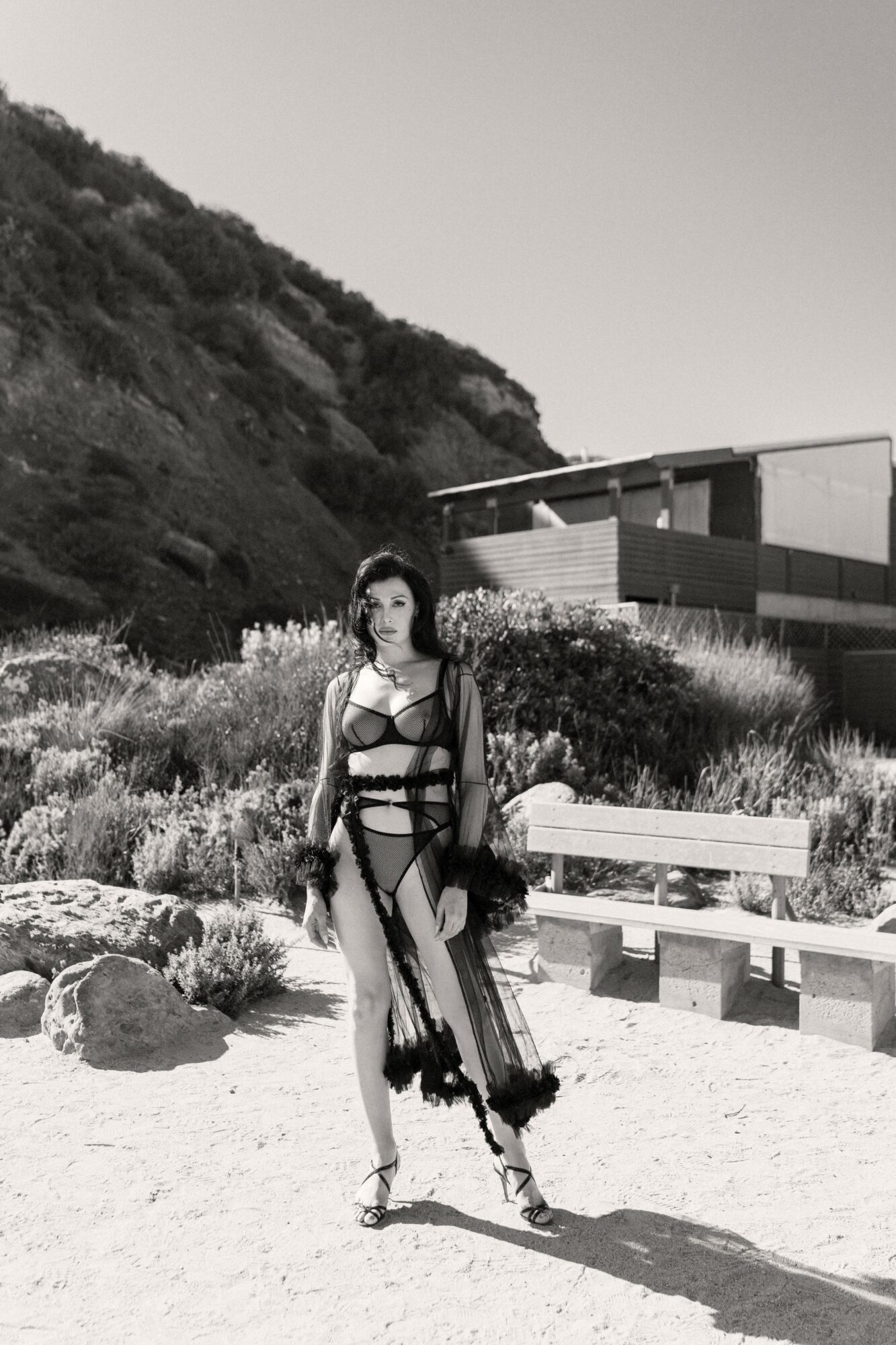 Woman in a dress standing on a sandy area with plants, rocks, and a modern building in the background.