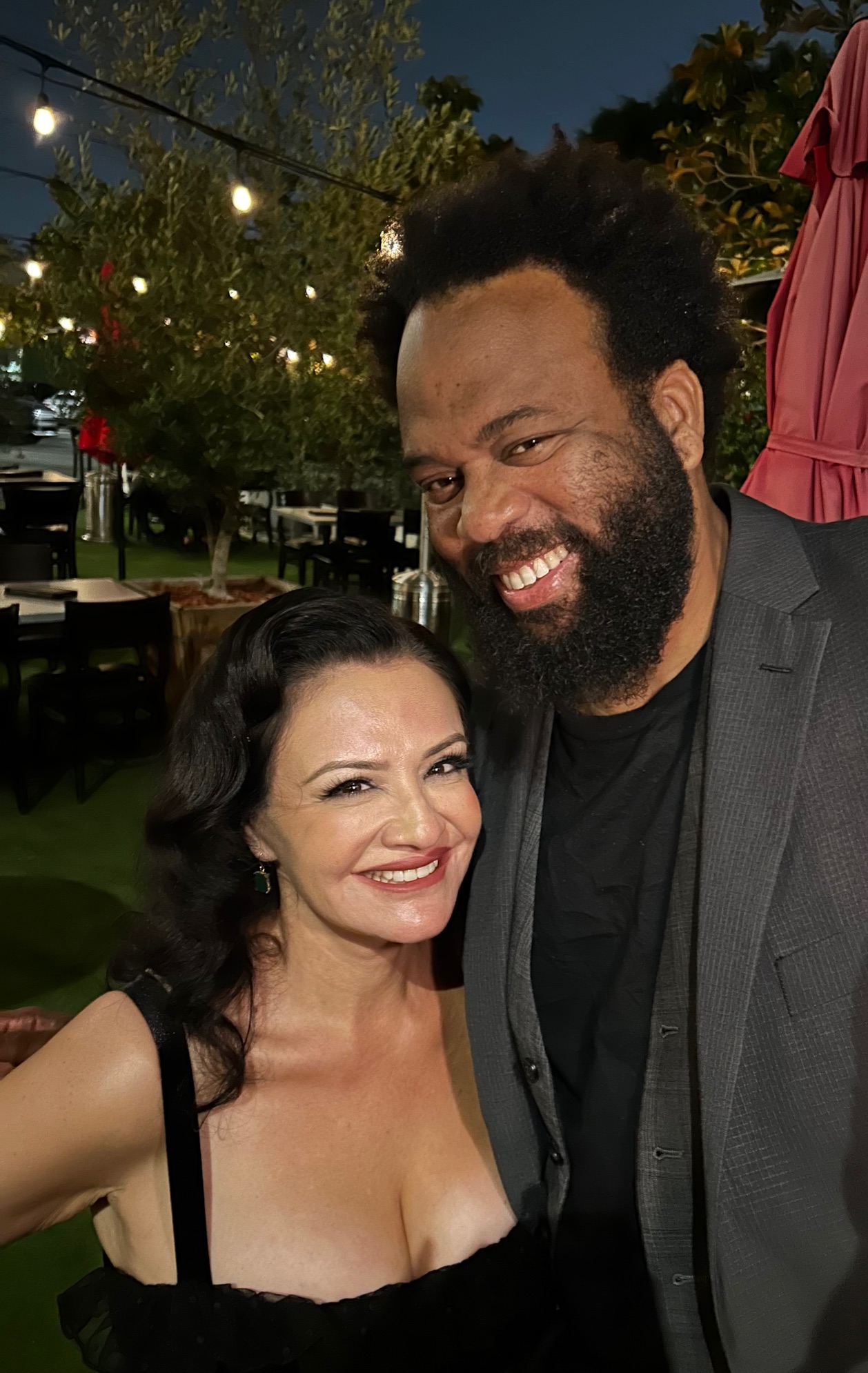 A woman and a man smiling outdoors at night, with string lights and trees in the background.