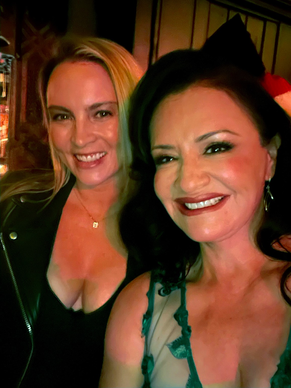 Two women smiling, one with blonde hair and the other with dark hair, in a dimly lit setting.