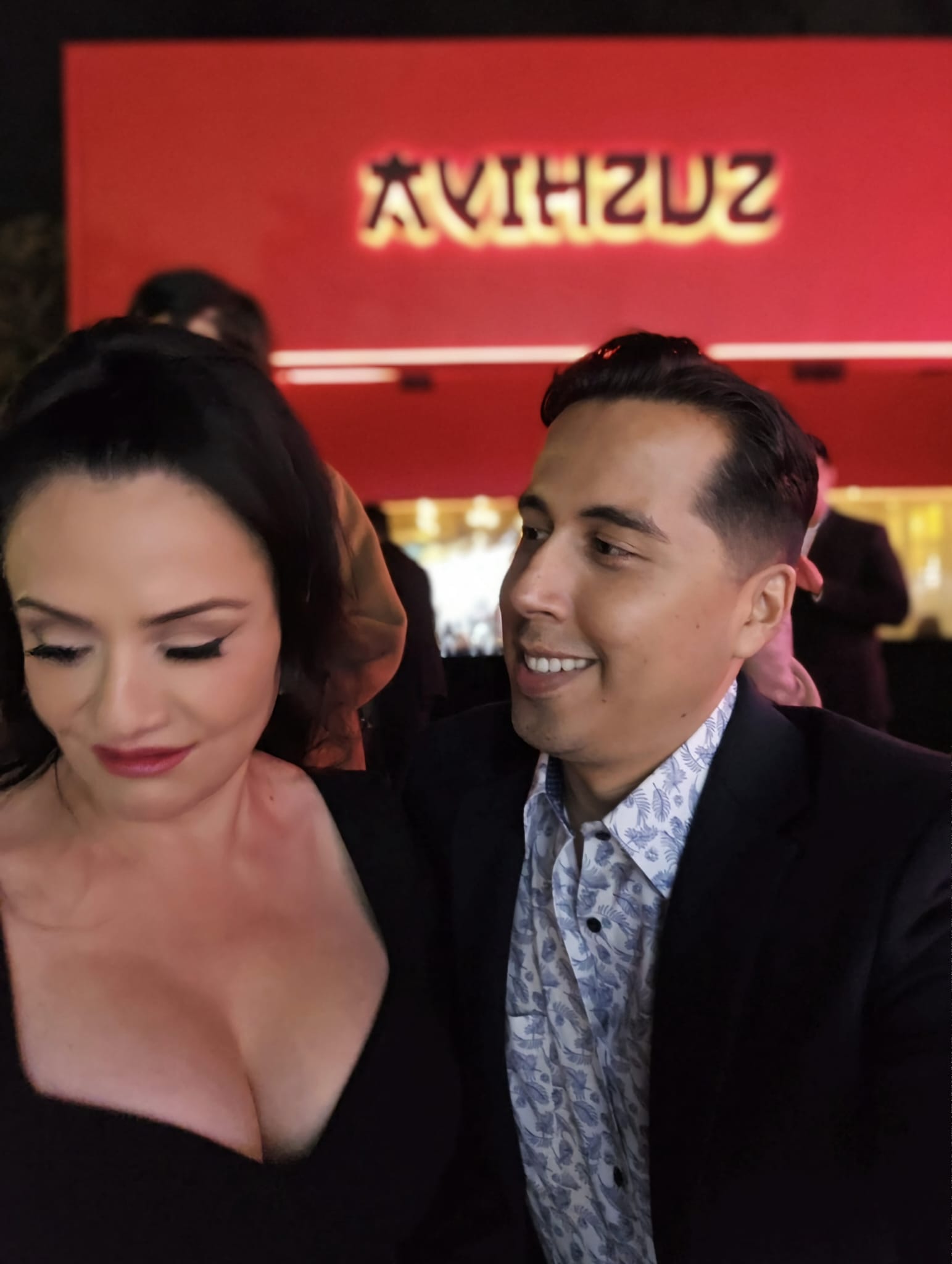 Two people smiling and looking at each other in a restaurant with a red wall and illuminated sign.