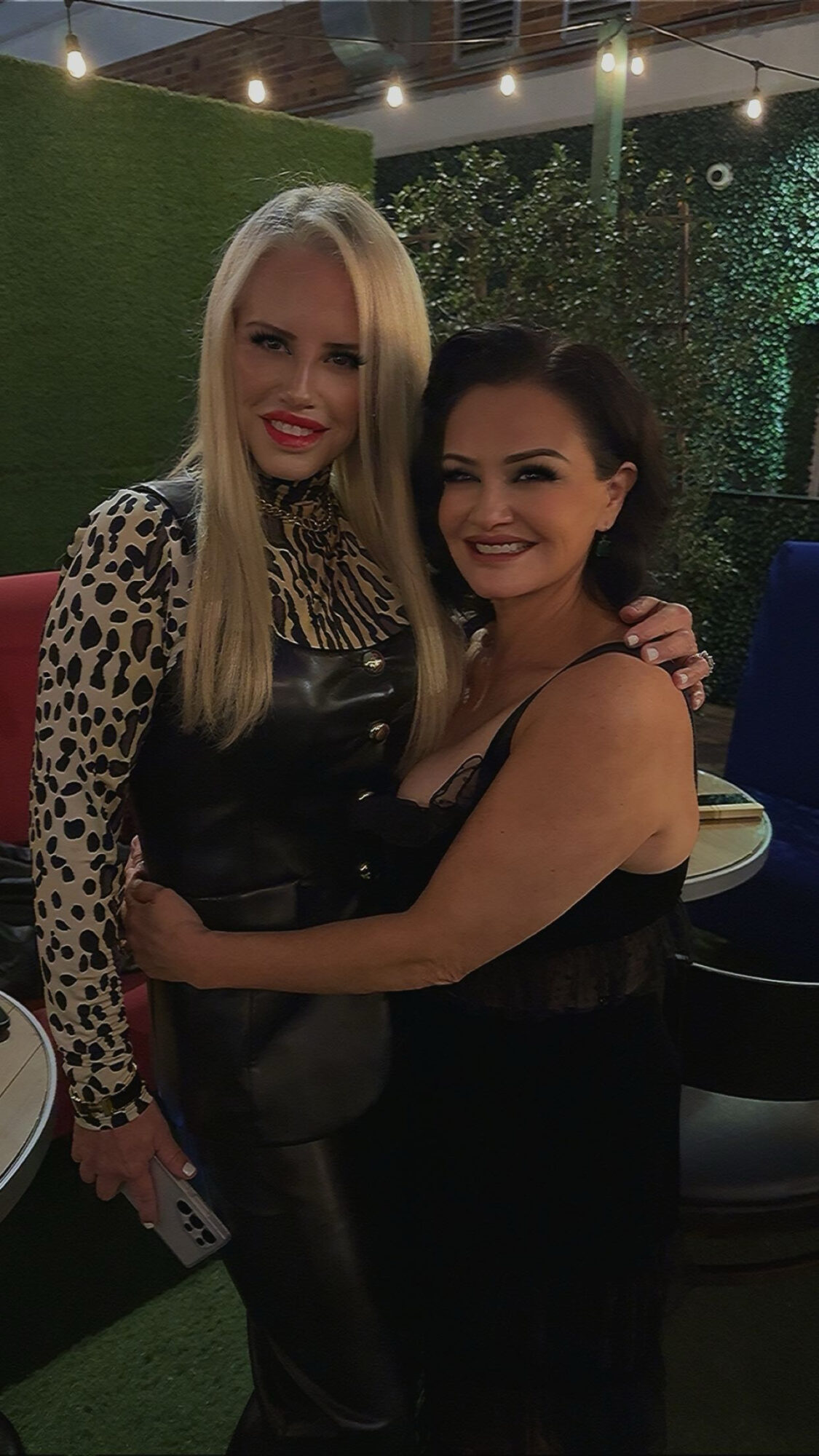 Two women smiling and hugging in an outdoor setting at night, with greenery and string lights in the background.