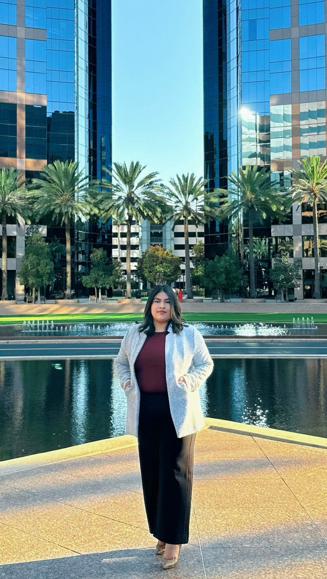 Woman standing outdoors in front of a water feature and modern glass buildings, palm trees behind her.