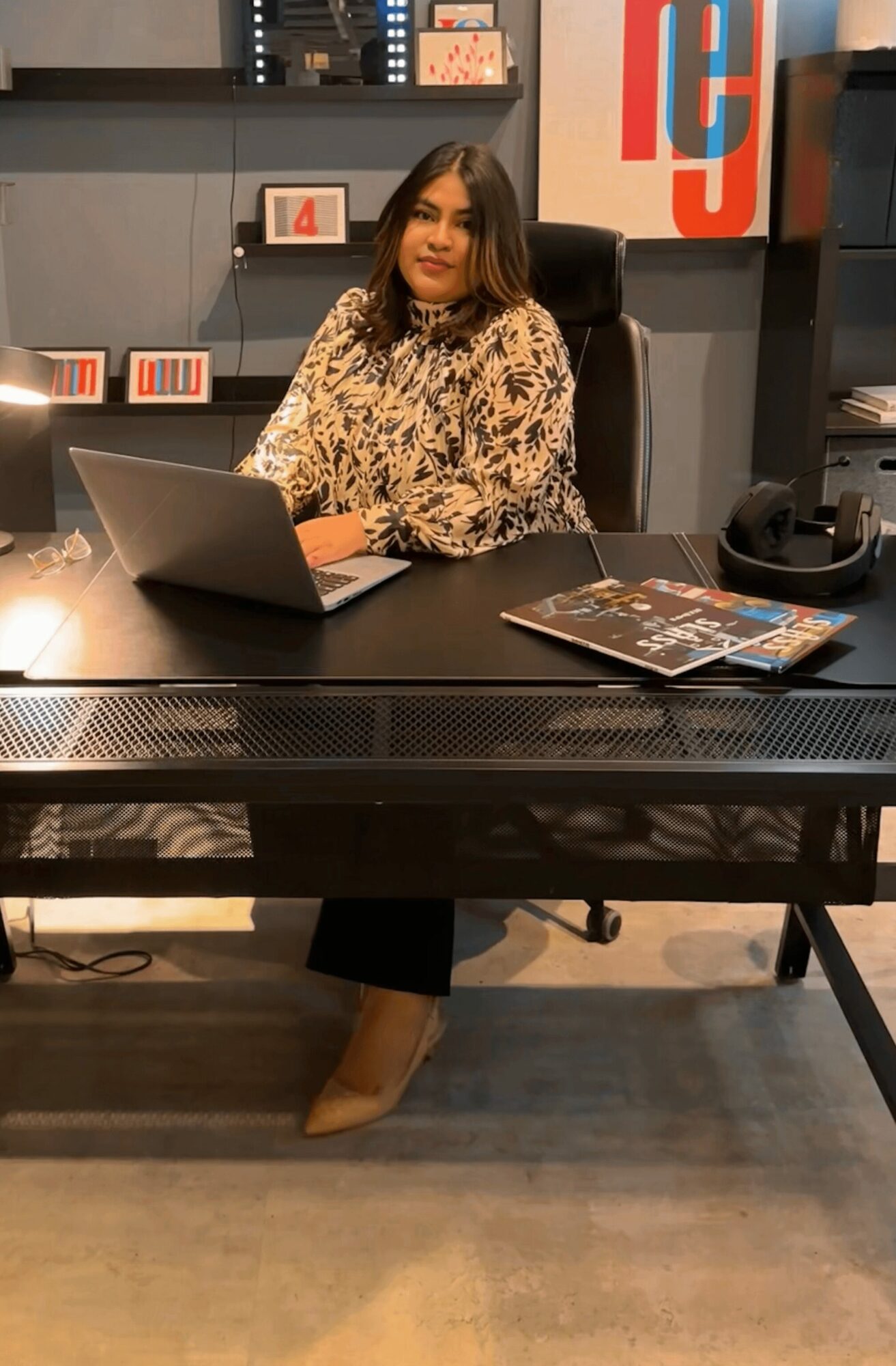 Woman sitting at desk with laptop, magazine, and headphones in office setting.