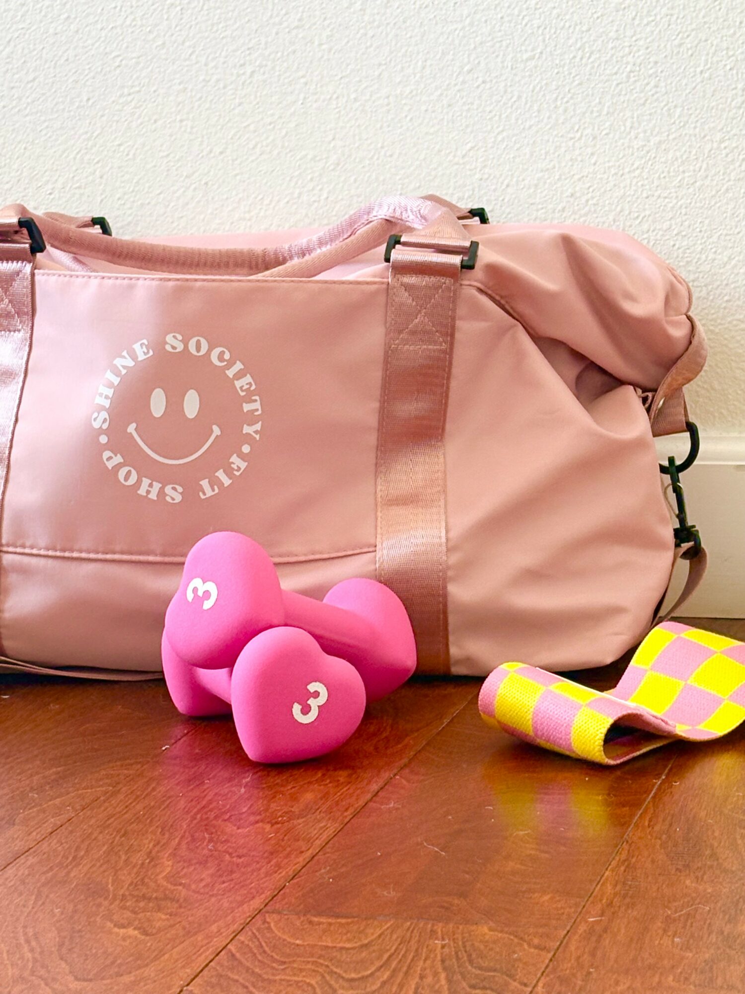 Pink gym bag with white logo, two pink dumbbells with number 3, and a yellow and pink checkered towel on a wooden floor.