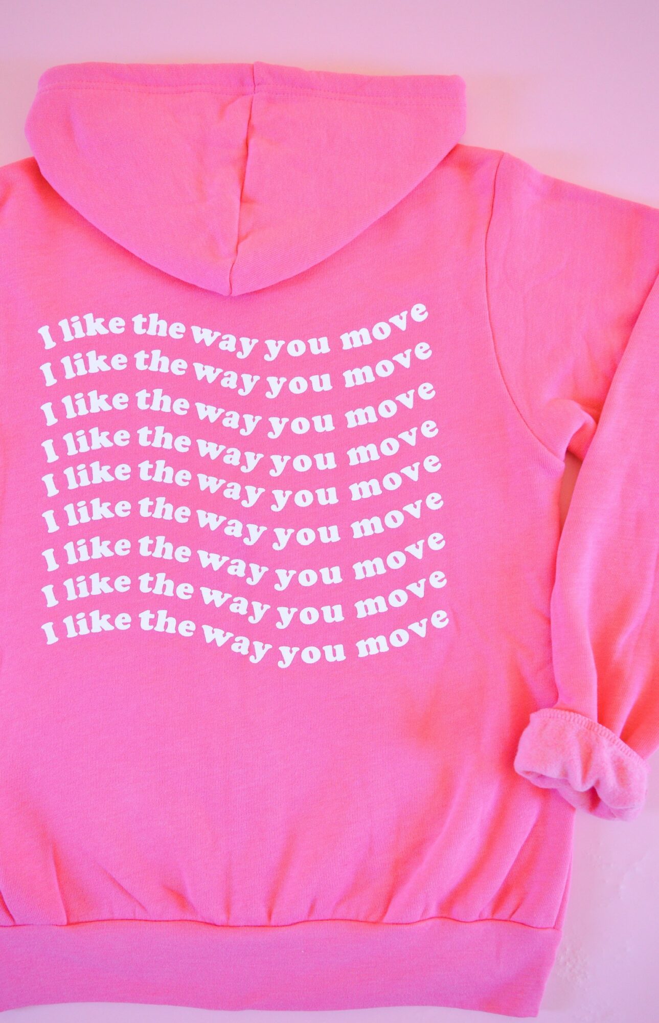 Pink hoodie with white text repeating 'I like the way you move' on the back, with a rolled-up sleeve.
