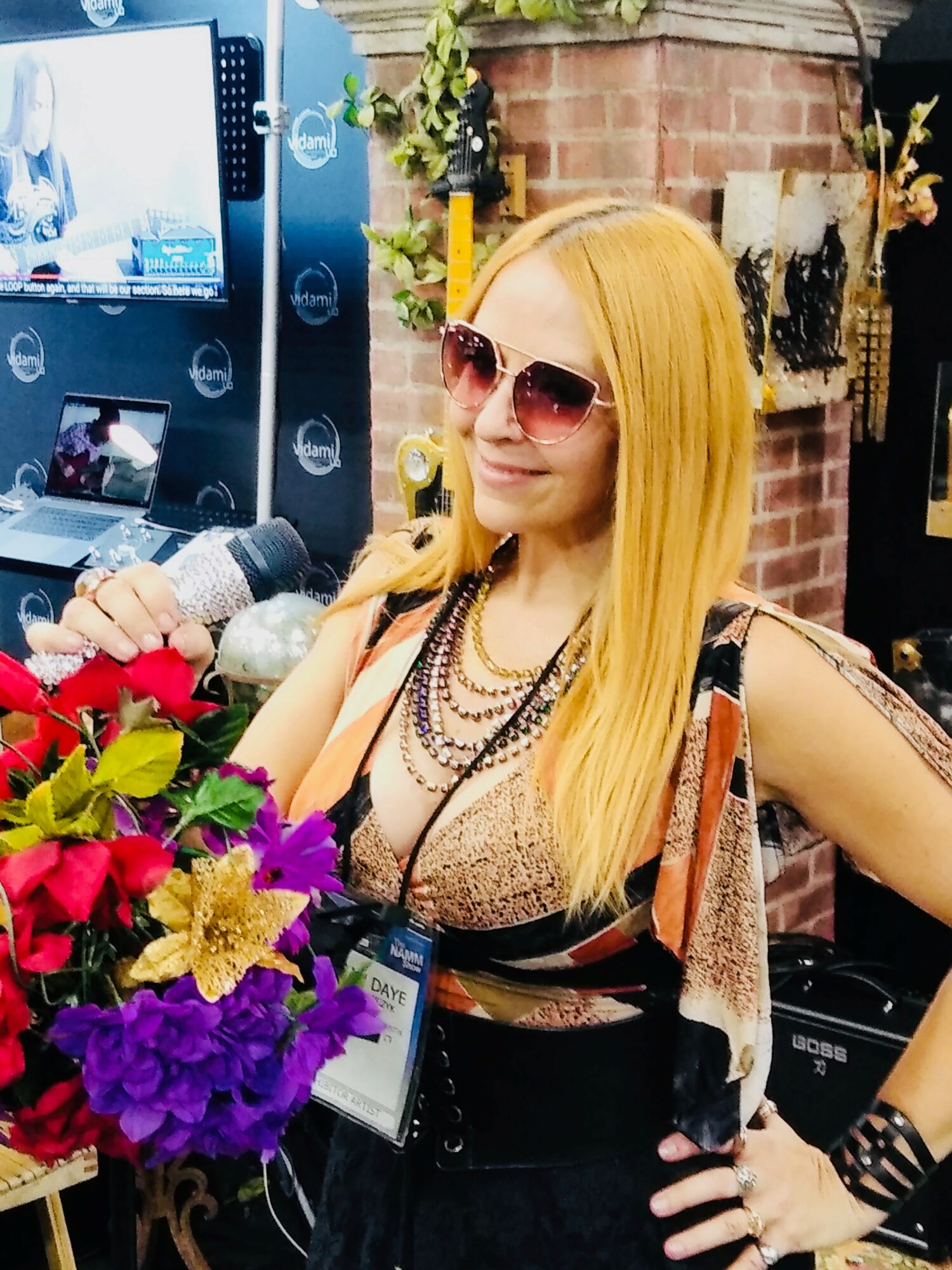 Woman with long blonde hair wearing sunglasses and layered necklaces, standing near a colorful flower arrangement indoors.