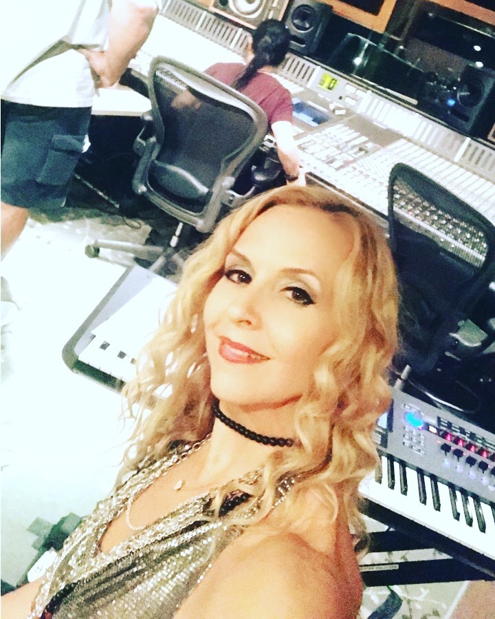 Woman with blonde curly hair taking a selfie in a music studio with mixing equipment and people working in the background.