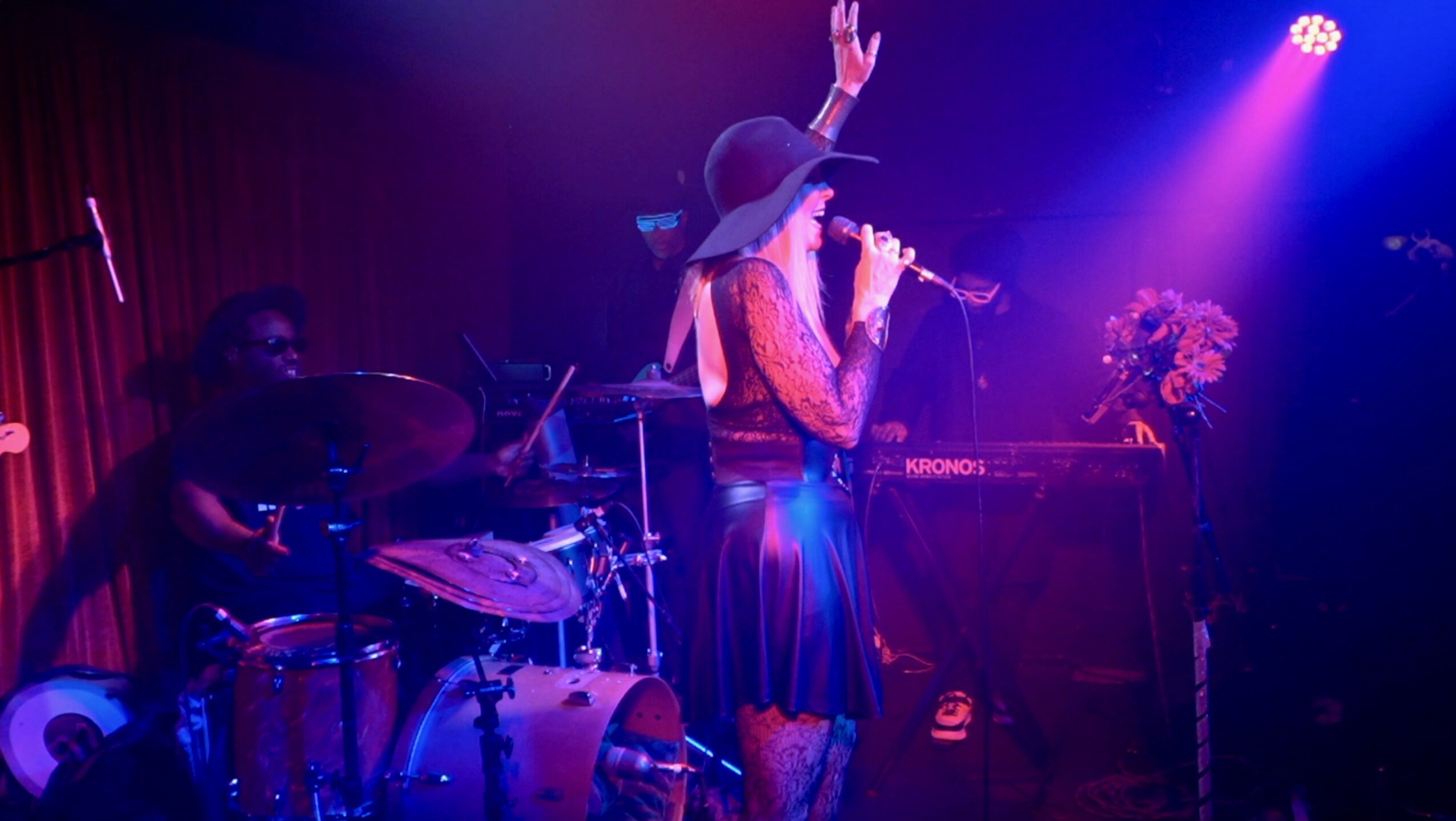 Performer singing into microphone with a band on stage, purple and blue lighting, and a flower arrangement nearby.