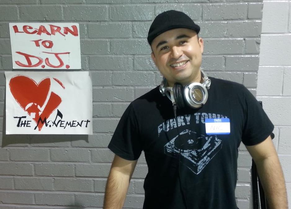 Smiling person wearing a black cap and headphones, standing in front of a brick wall with a sign.