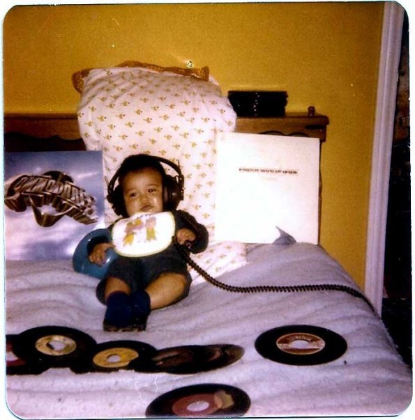Child with headphones lying on bed, holding a record, surrounded by records, pillow, and yellow wall background.