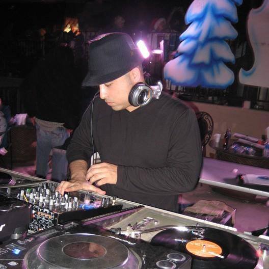 DJ wearing a black hat and headphones, operating a turntable and mixer in a dimly lit club or event space.