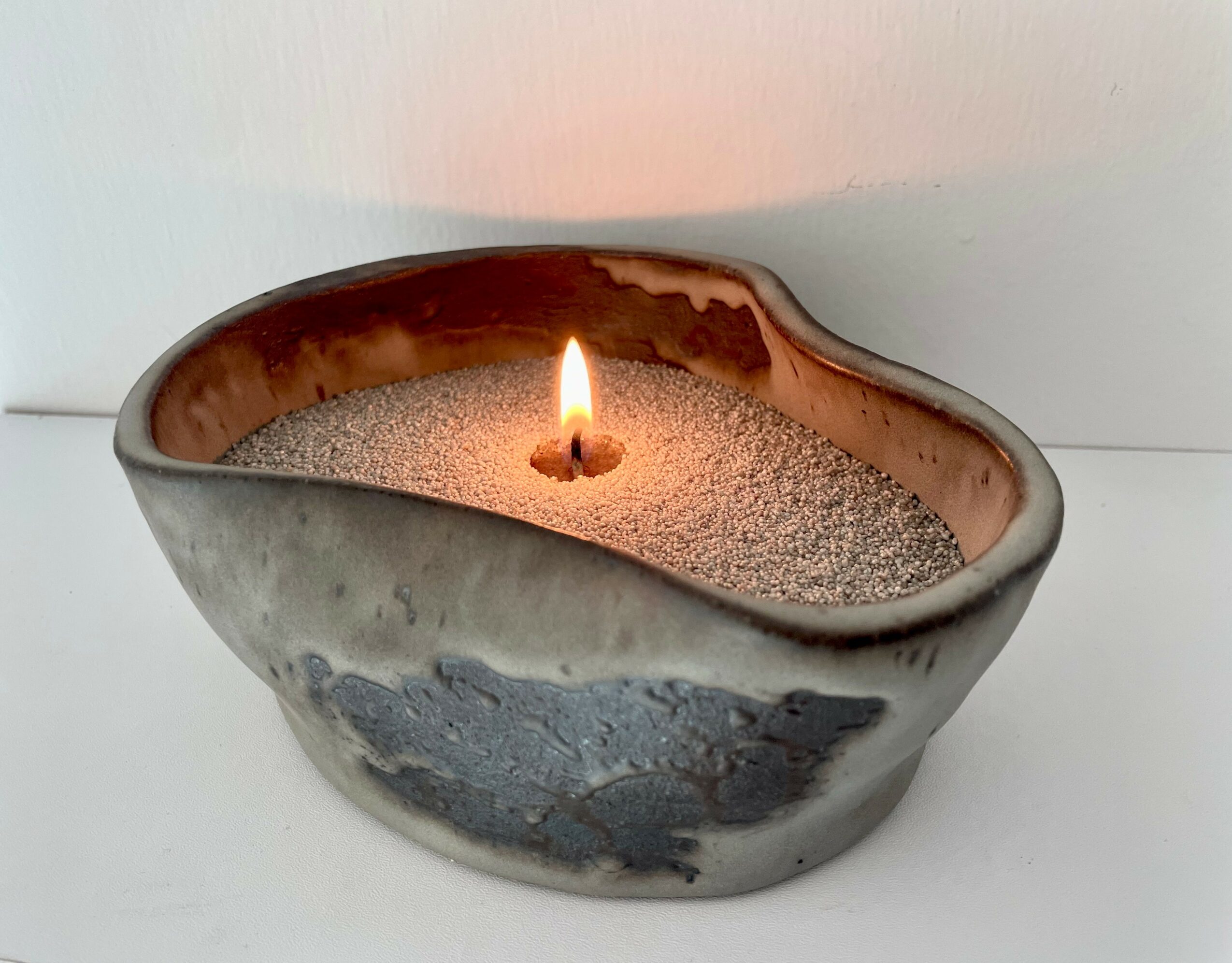 Ceramic bowl filled with sand and a lit candle, placed on a white surface.