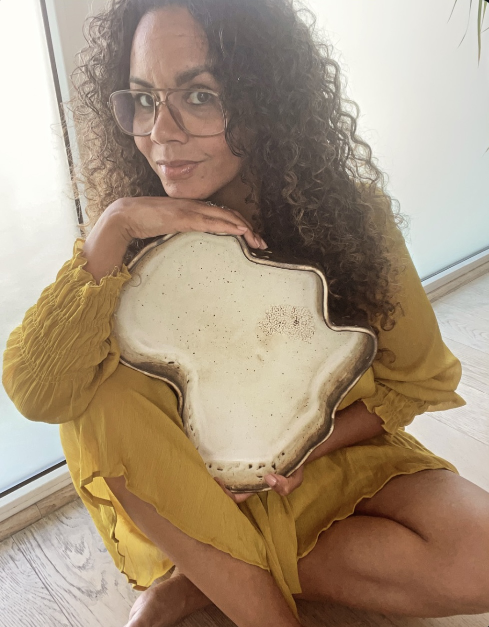 Woman with curly hair and glasses sitting on floor, holding a large, irregularly shaped object, wearing a yellow dress.