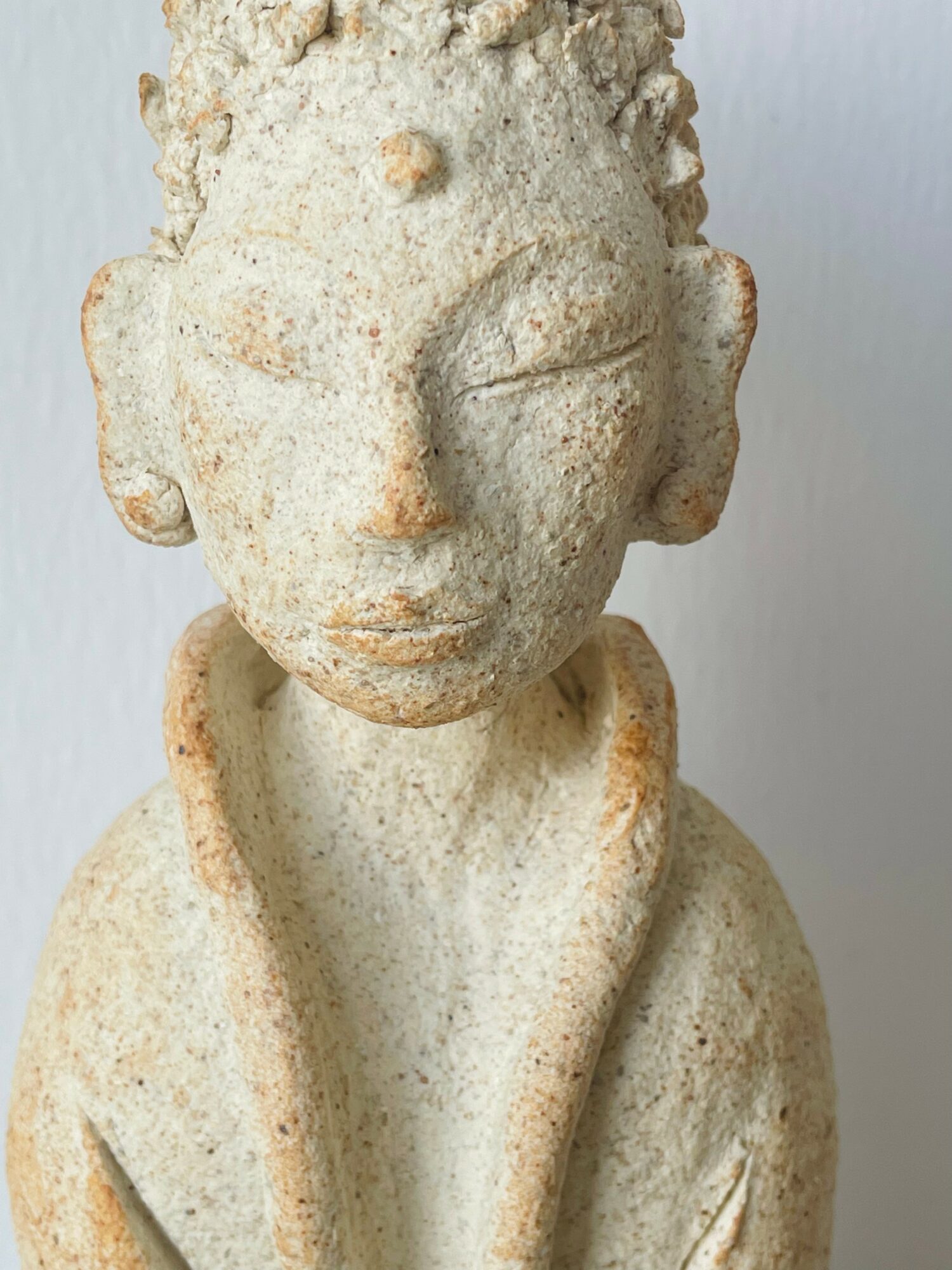 Stone sculpture of a person with closed eyes and textured hair, wearing a robe or cloak, against a plain background.
