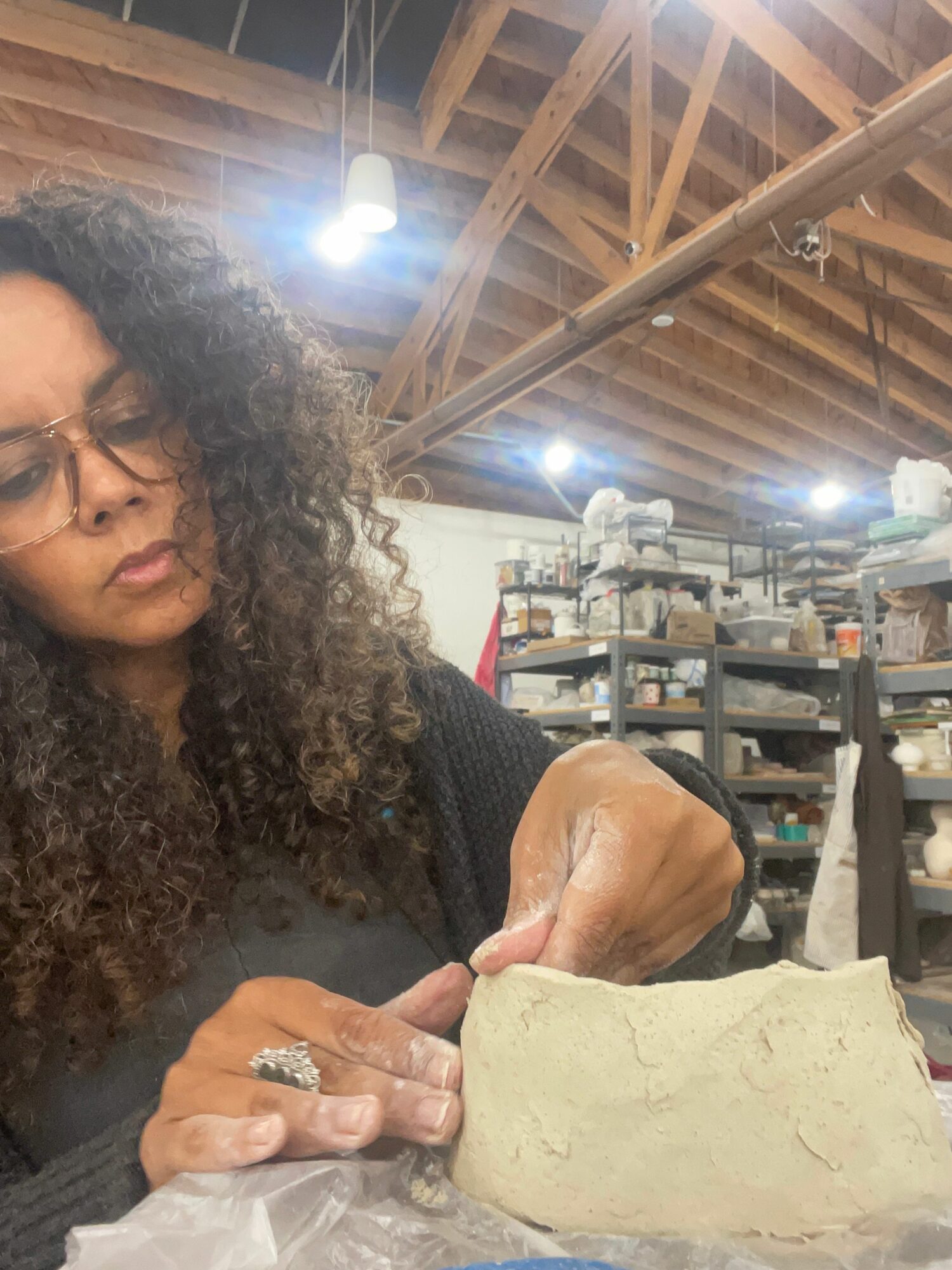 Woman with curly hair and glasses working with clay in a workshop with shelves and tools in background.