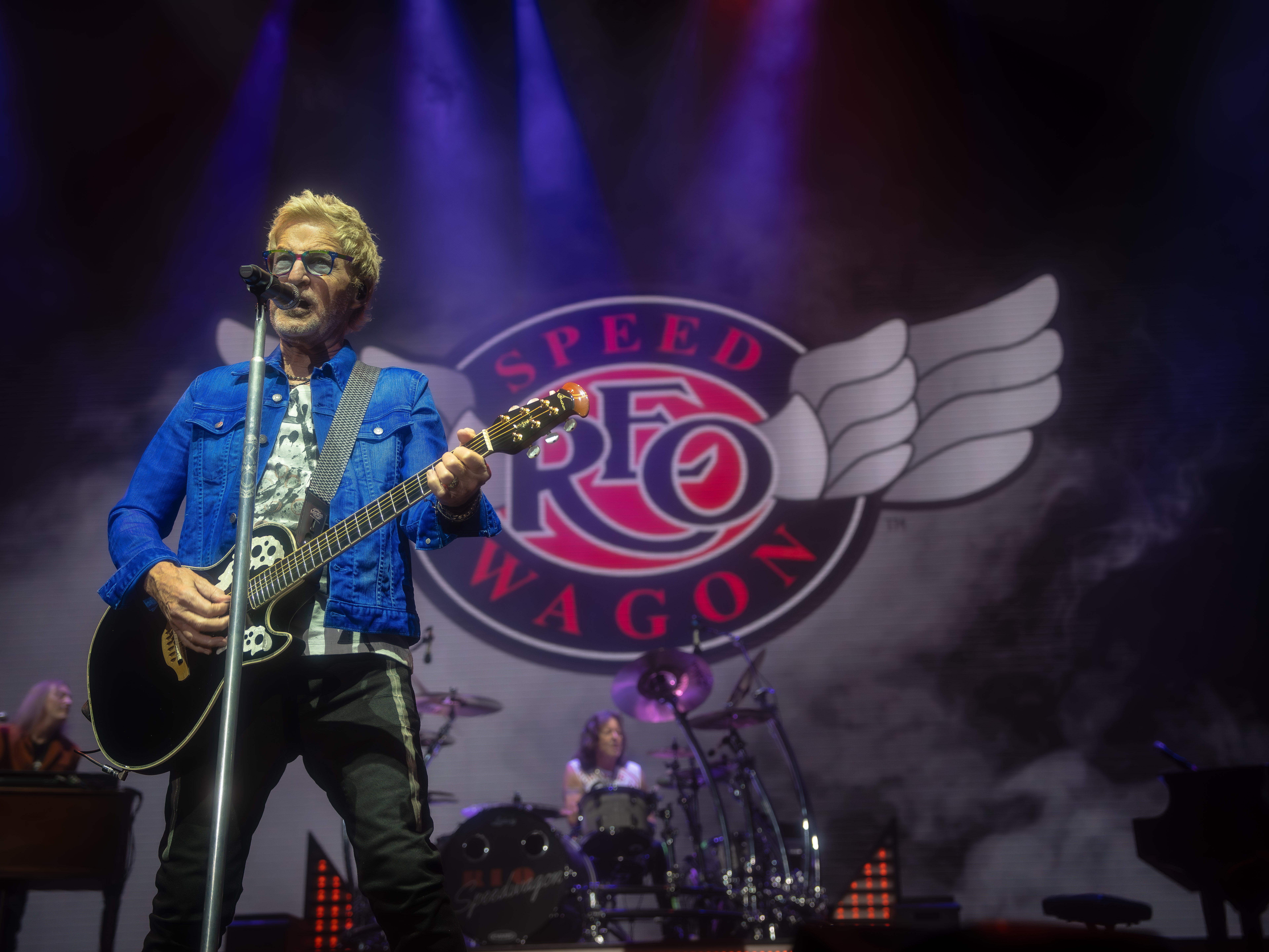 Musician with sunglasses and blue jacket playing guitar on stage with a drum set in the background.