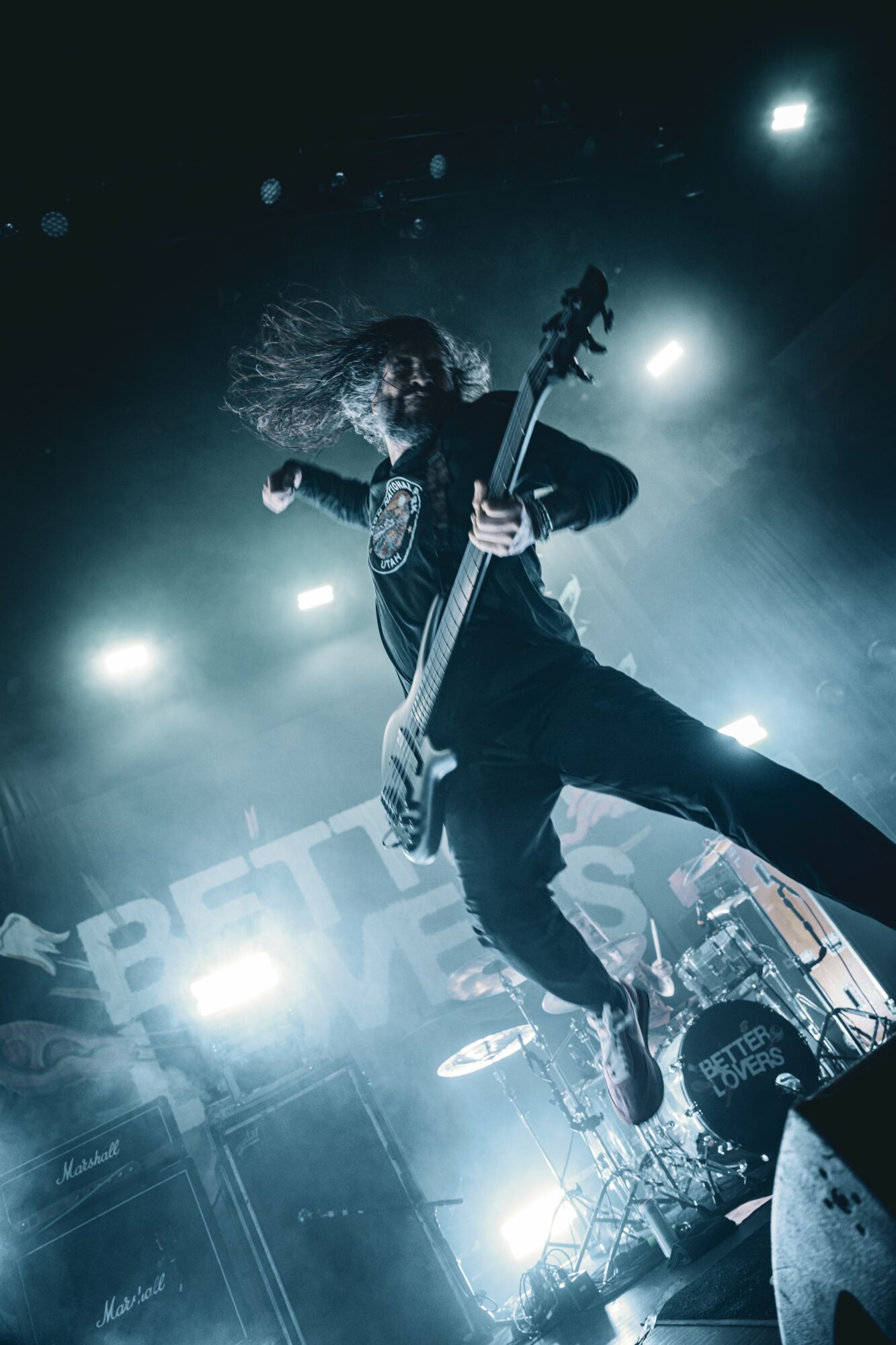 Musician jumps in air playing bass guitar on stage with bright lights and equipment around.