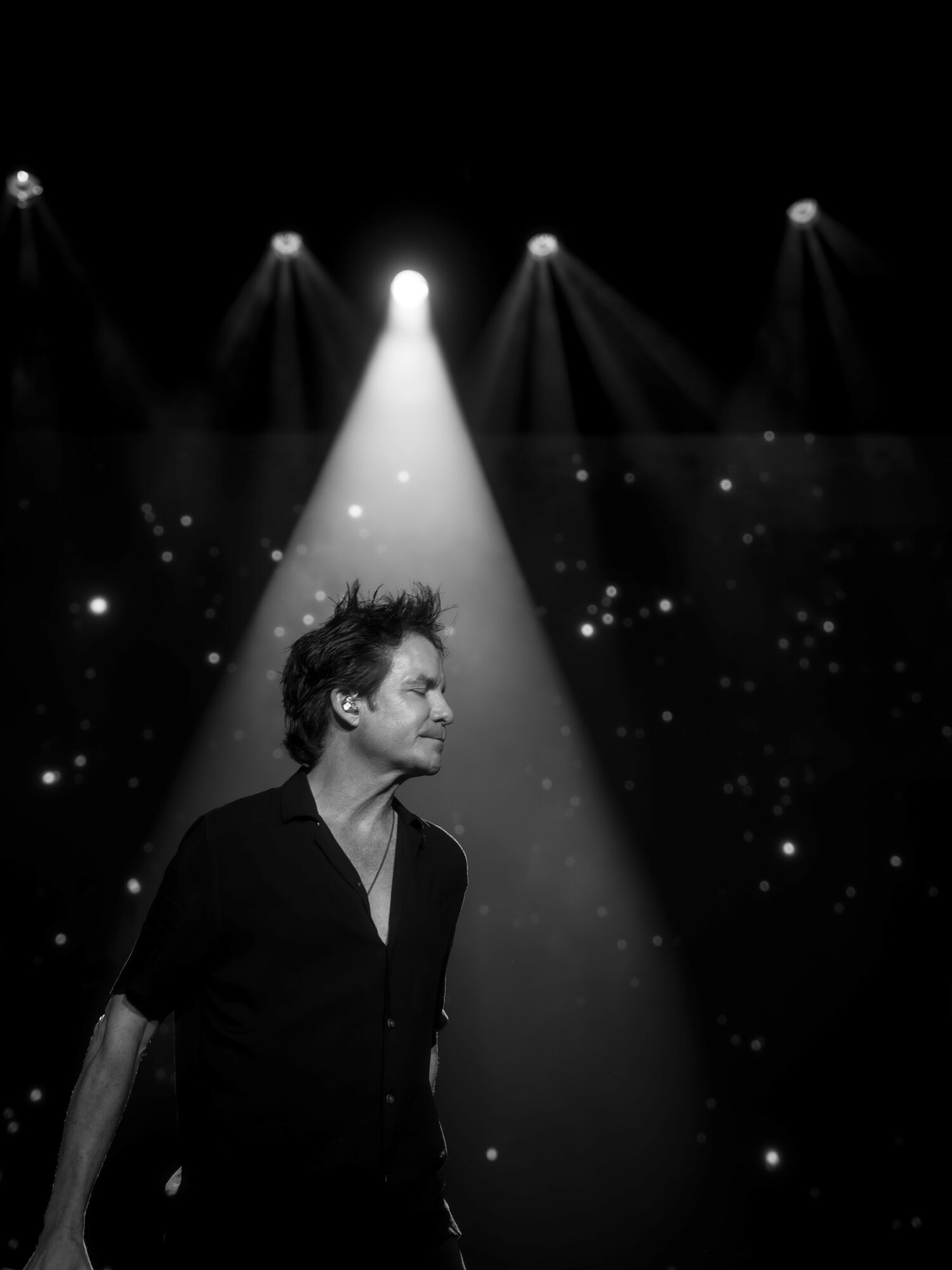 Person with short hair and dark shirt standing on stage with spotlight and starry background.