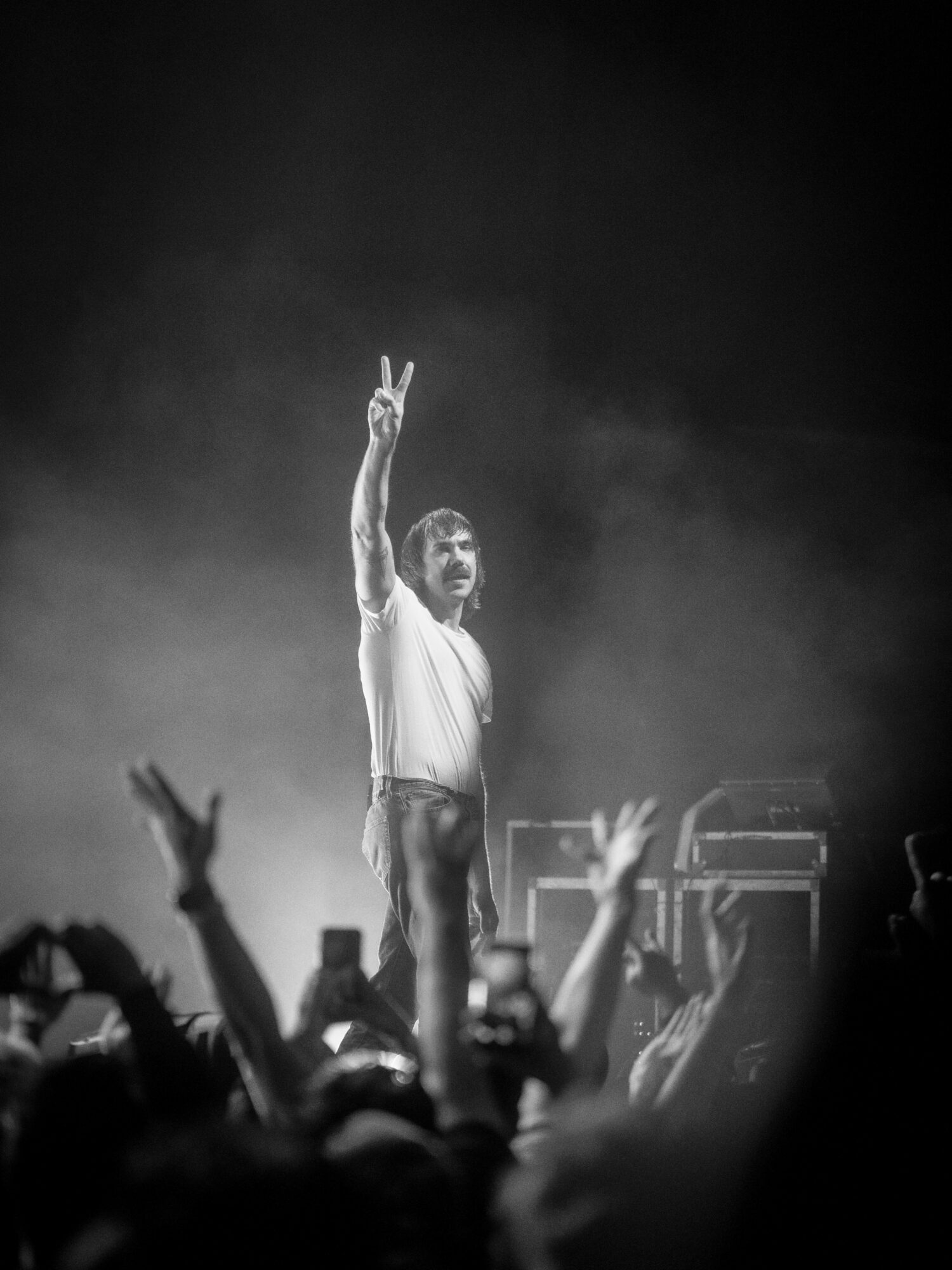 Performer on stage making a peace sign with one hand, audience raising hands, black background.