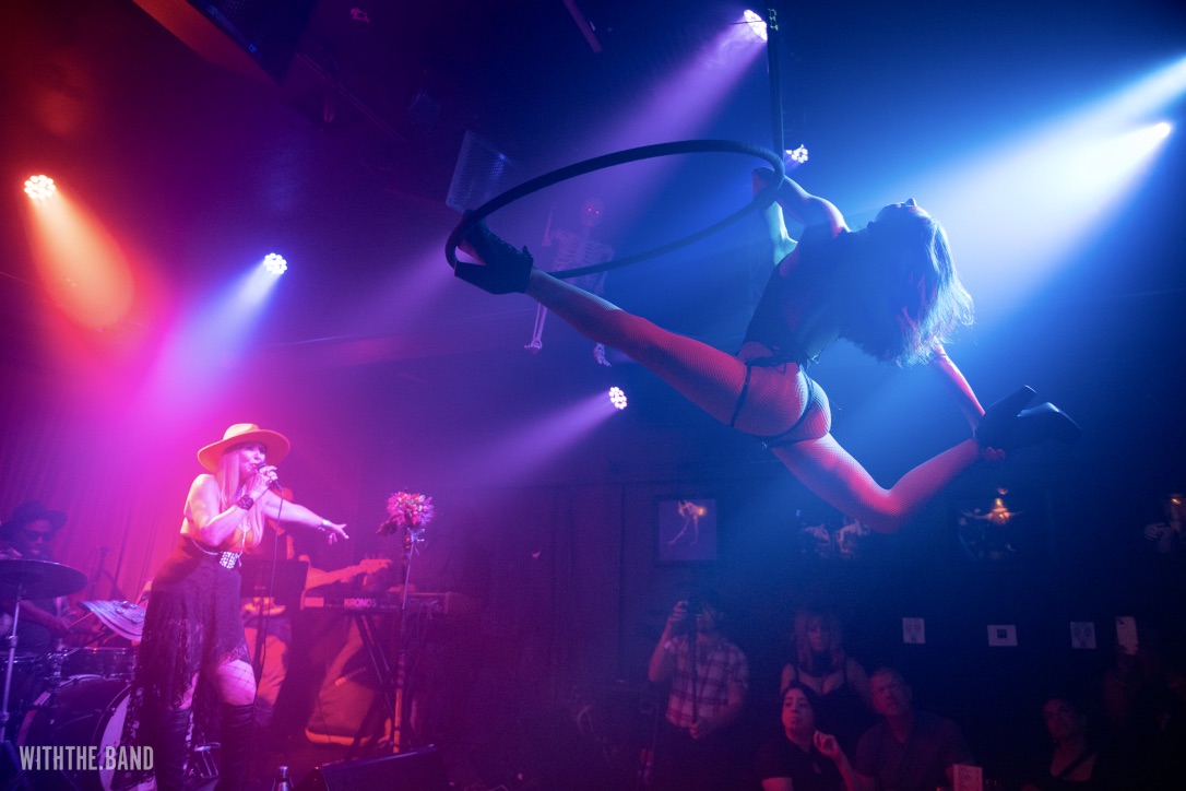 A performer is suspended in the air with a hoop, illuminated by colorful stage lights, in a lively concert setting.