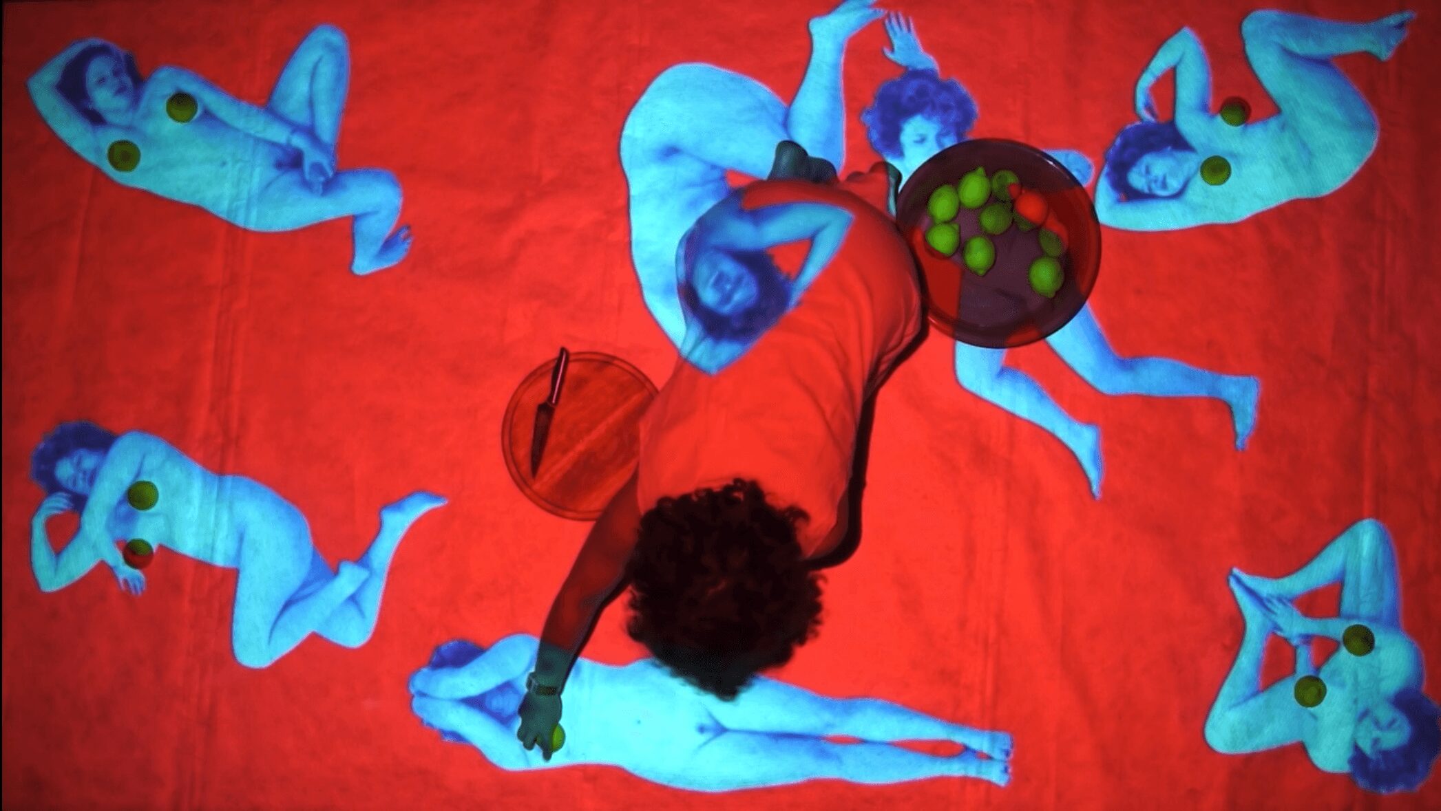 Person lying on red surface surrounded by six blue plush animal toys, holding a bowl of green grapes.