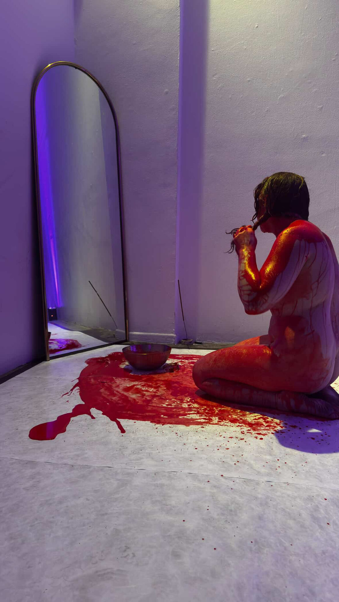 Person sitting on floor with legs crossed, holding a cup, near a mirror and a bowl, with red paint on floor.