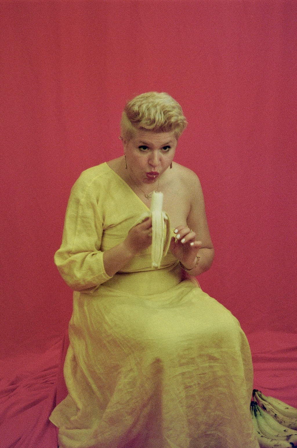 Woman in yellow dress sitting on pink background, holding and eating a banana, with a surprised expression.