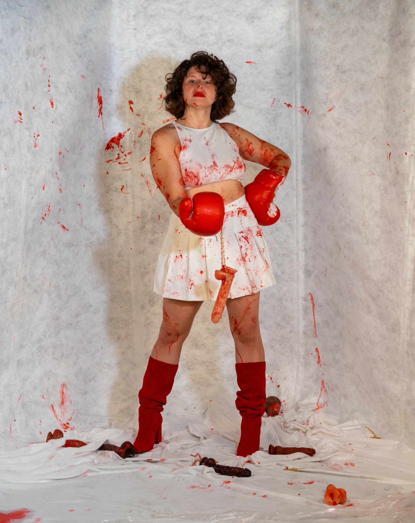 Woman with curly hair in a white dress with red paint, red boxing gloves, and red leg warmers, standing against a wall.