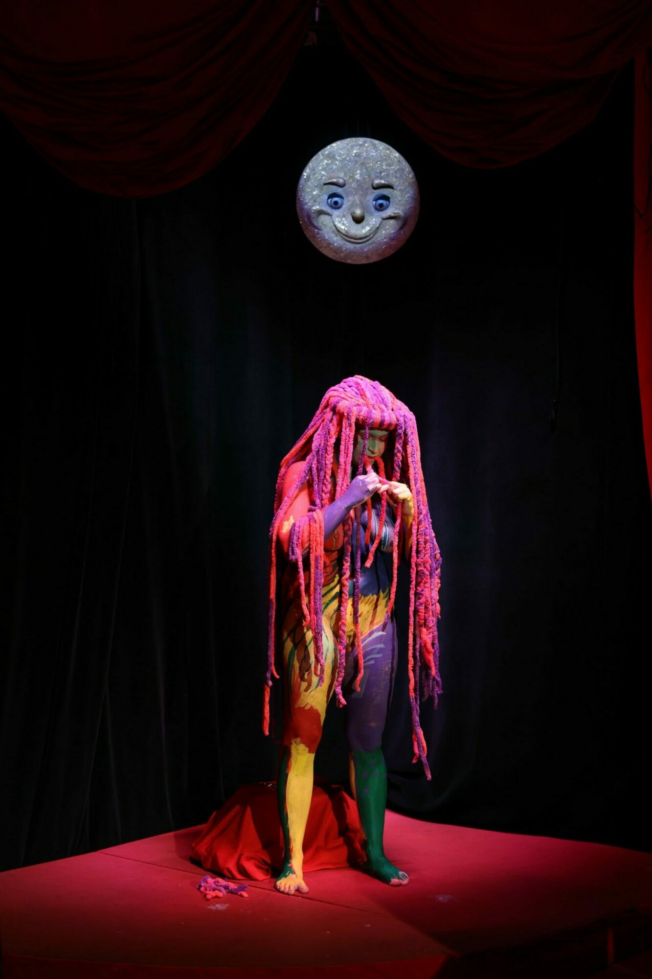 Person in colorful costume standing on stage with a moon prop in background, black curtain backdrop.