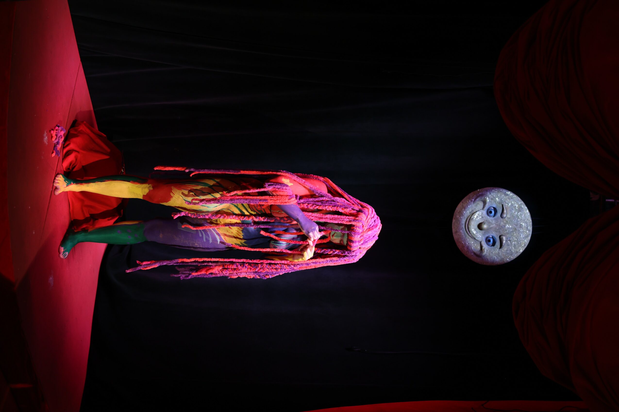 Person in colorful costume standing on stage with a moon prop in background, black curtain backdrop.