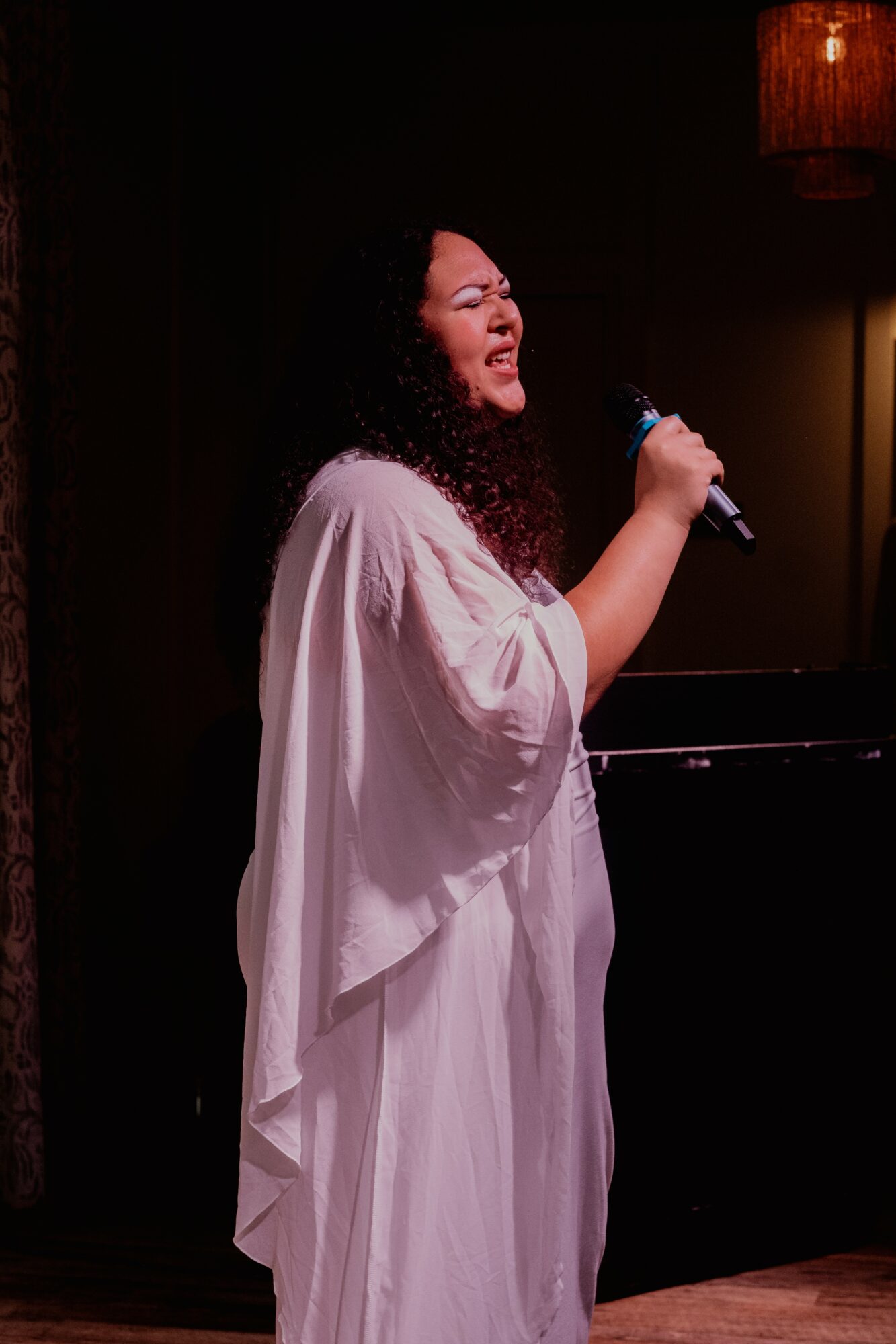 Woman singing into a microphone on stage with a dark background and warm lighting.