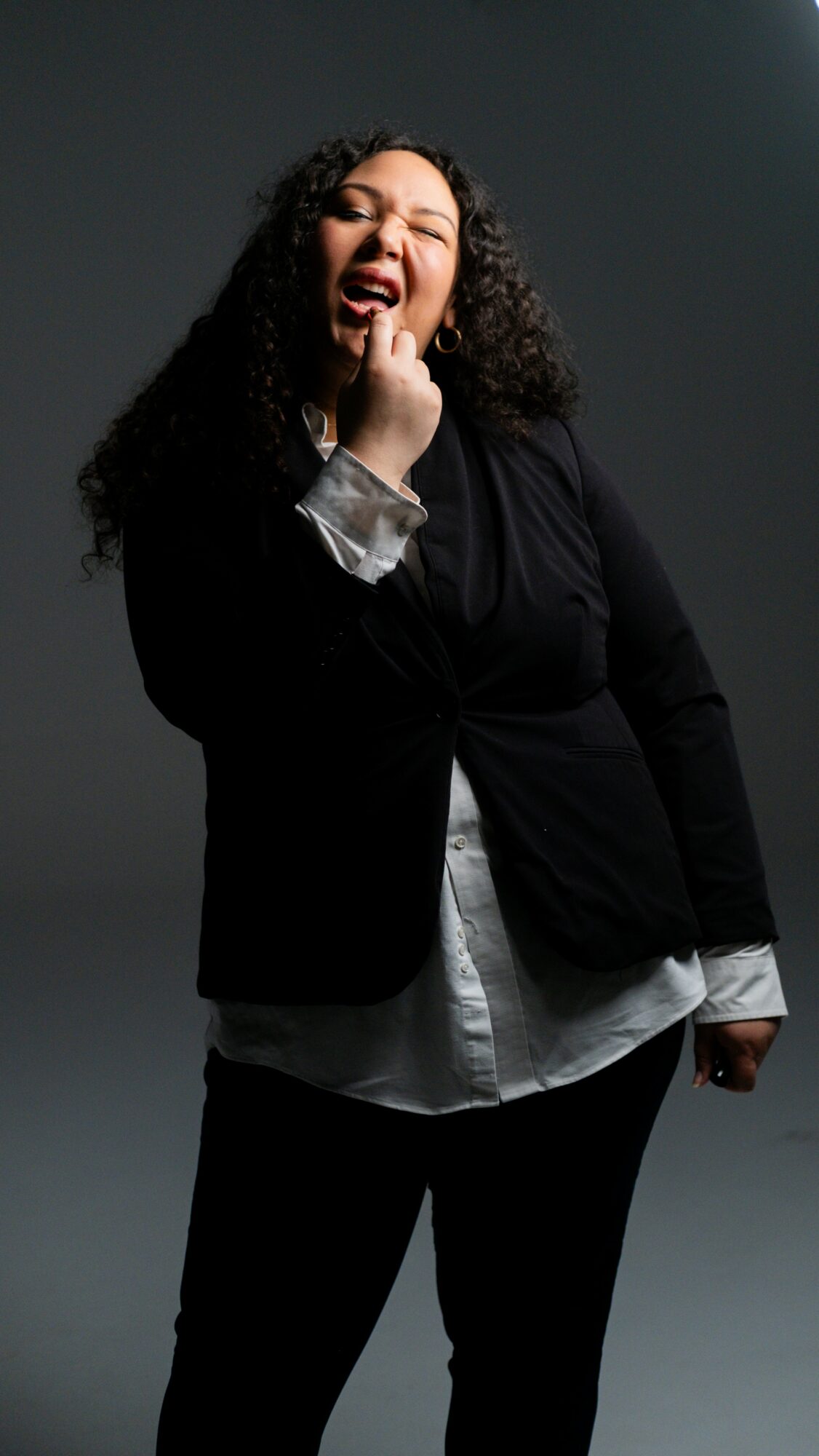 Woman with curly hair laughing, hand near mouth, wearing black jacket and white shirt, dark background.