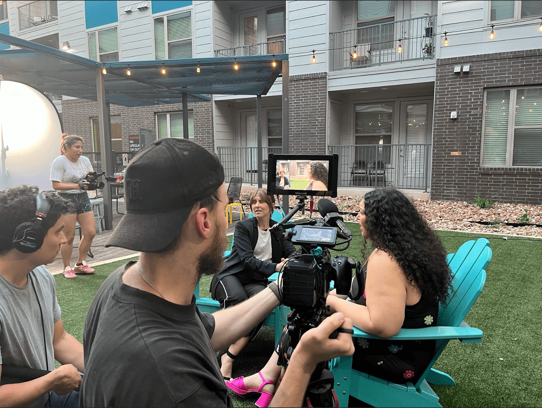 Filmmaker records interview with woman sitting on turquoise chair outdoors, with people and modern buildings in background.