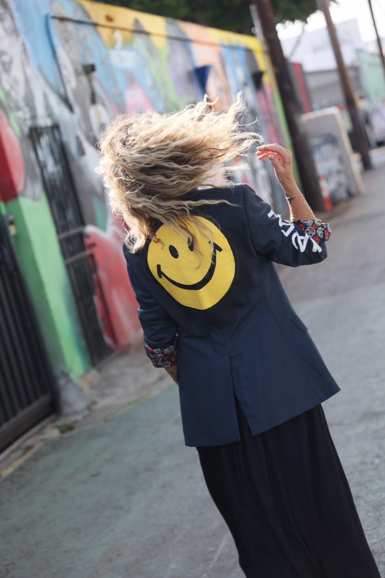 Person with curly hair wearing a navy coat with a yellow smiley face on the back, walking on a street with graffiti-covered wall.