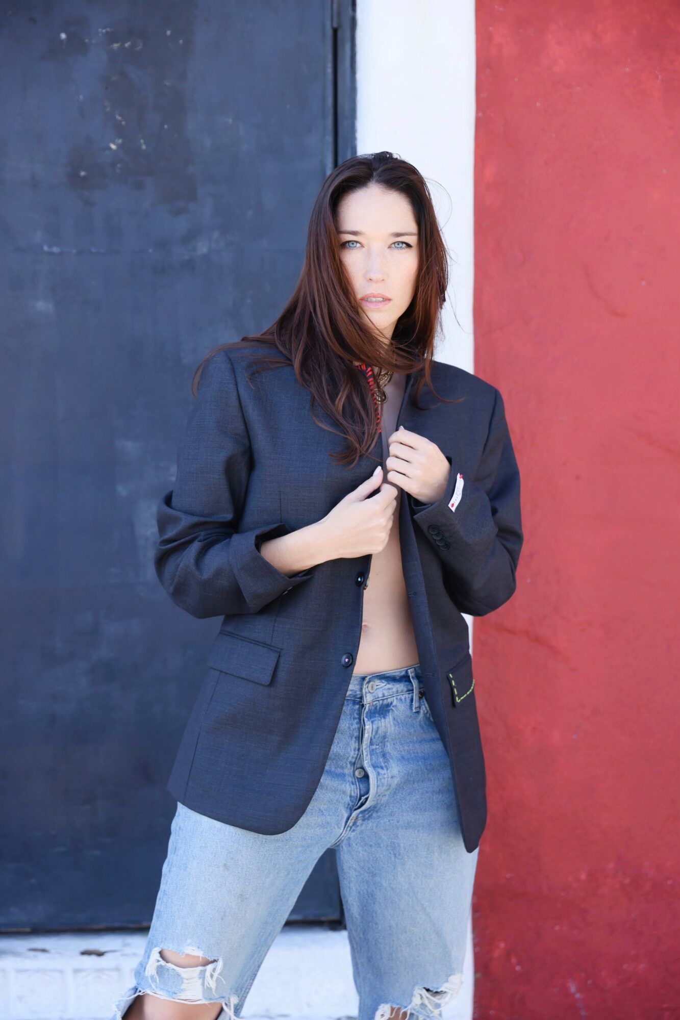 Woman with long dark hair wearing a blazer and ripped jeans standing against a colorful wall.