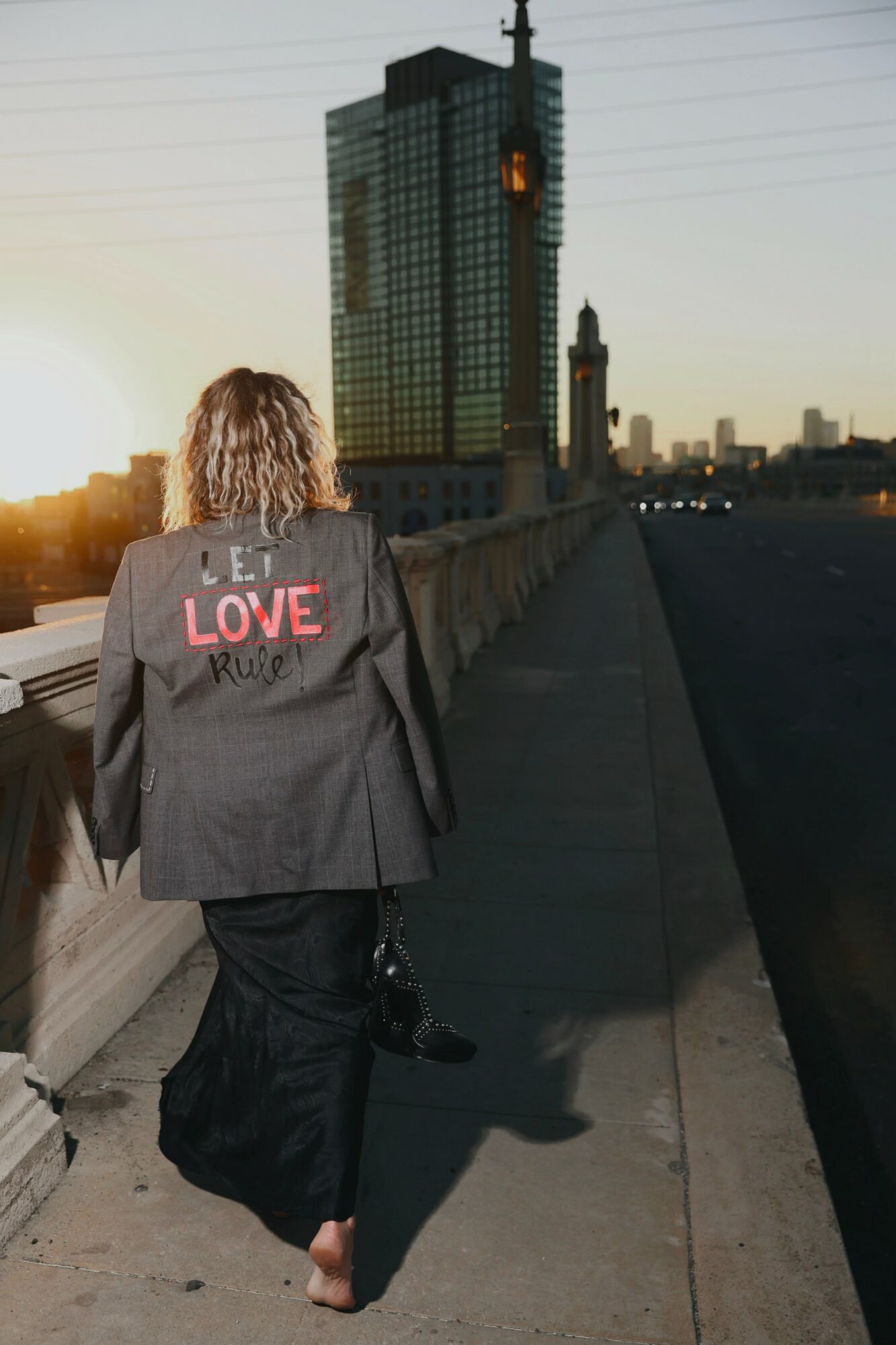 Person with curly hair walking on a bridge during sunset, city skyline with tall buildings in background.