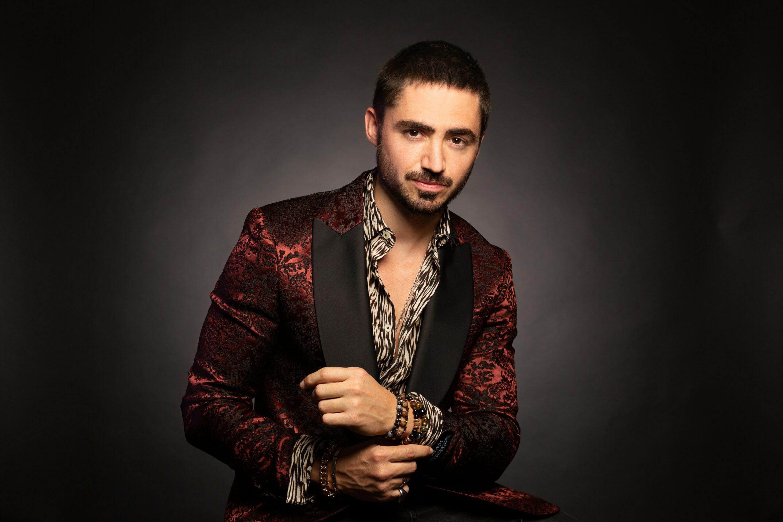 Man with dark hair and beard wearing a patterned shirt and a red and black blazer, posing against a dark background.