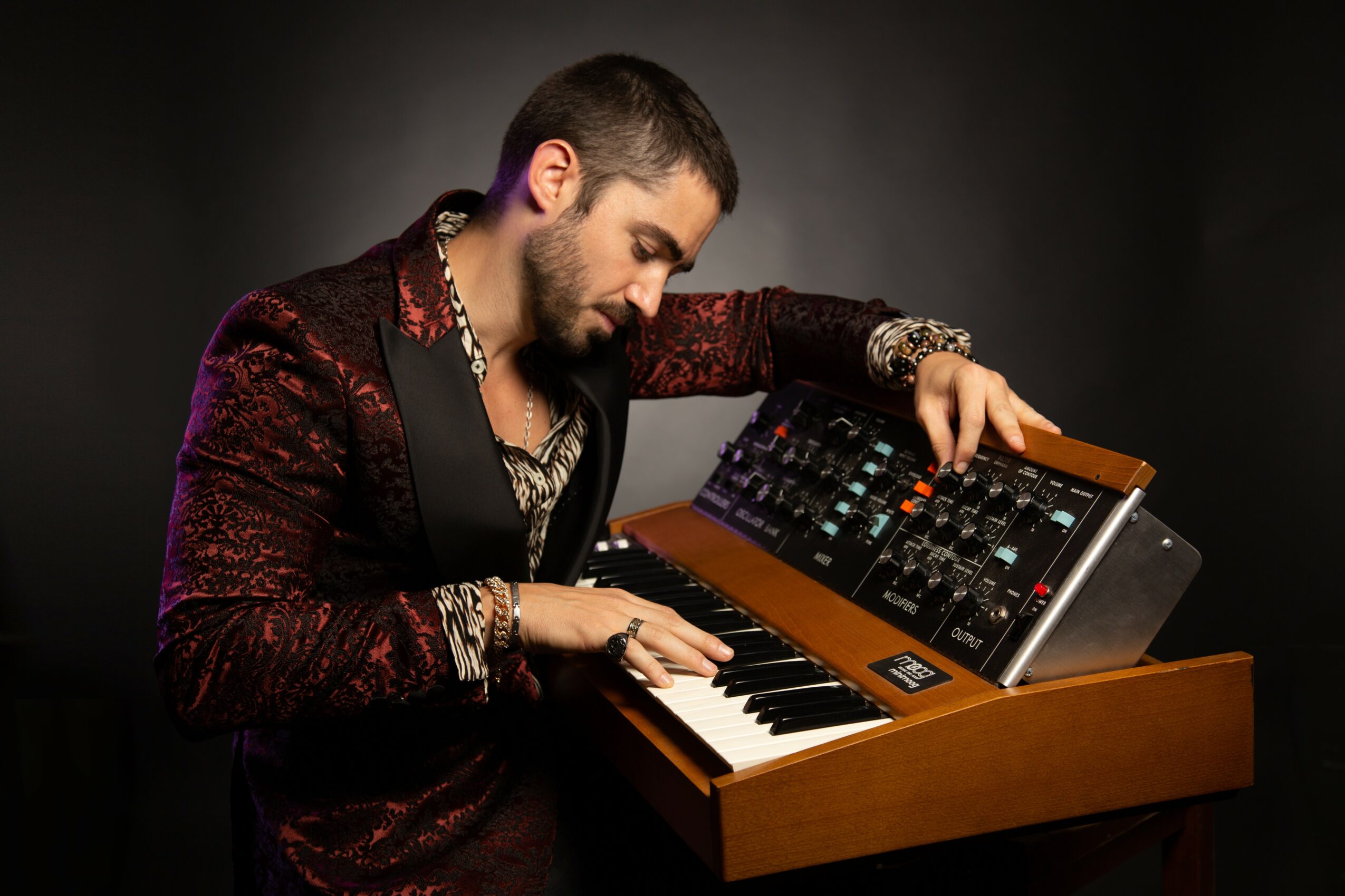 Man with beard and dark hair adjusts a synthesizer keyboard on a wooden stand, wearing a patterned blazer and jewelry.